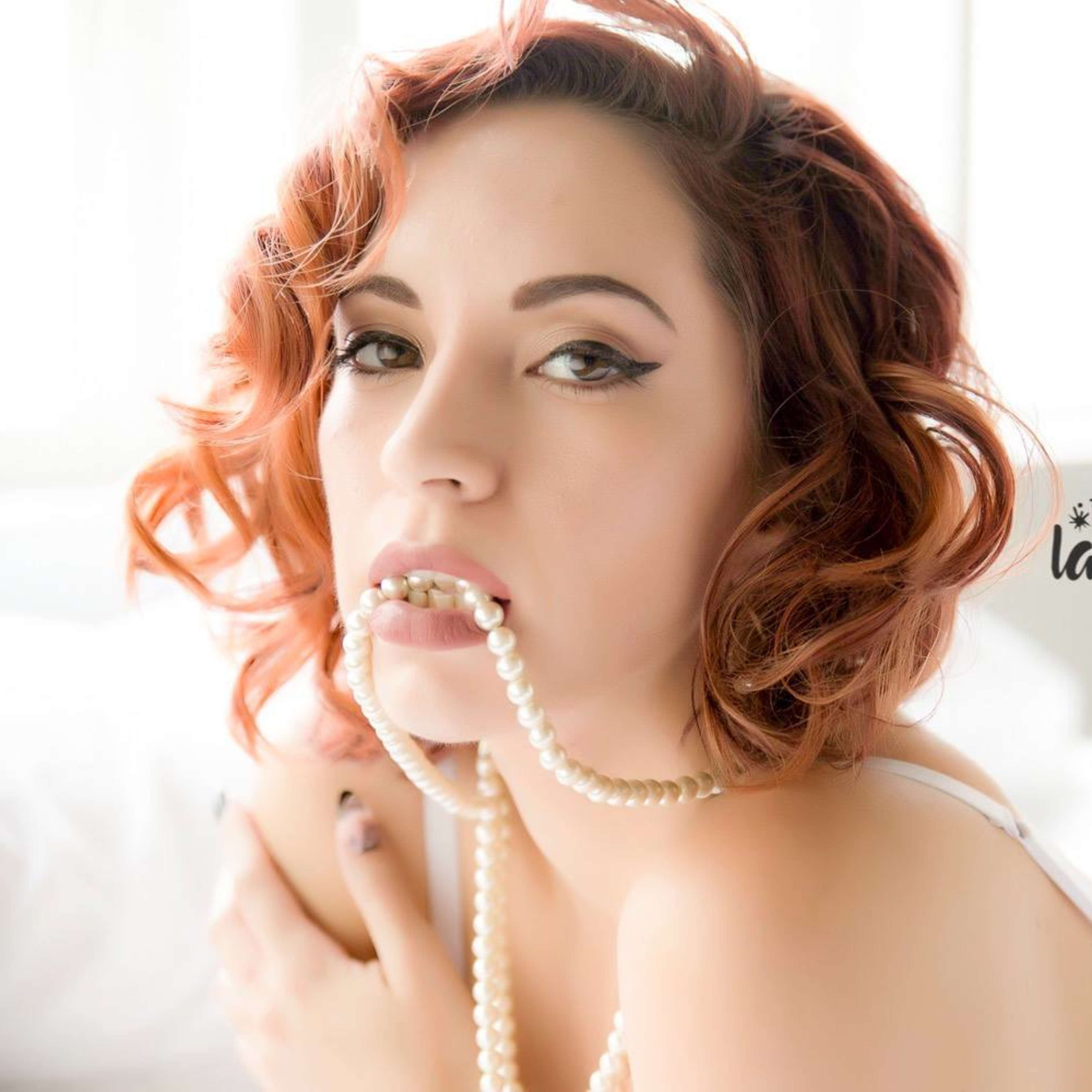A woman with curly red hair poses on a bed while holding a strand of pearls in her mouth.