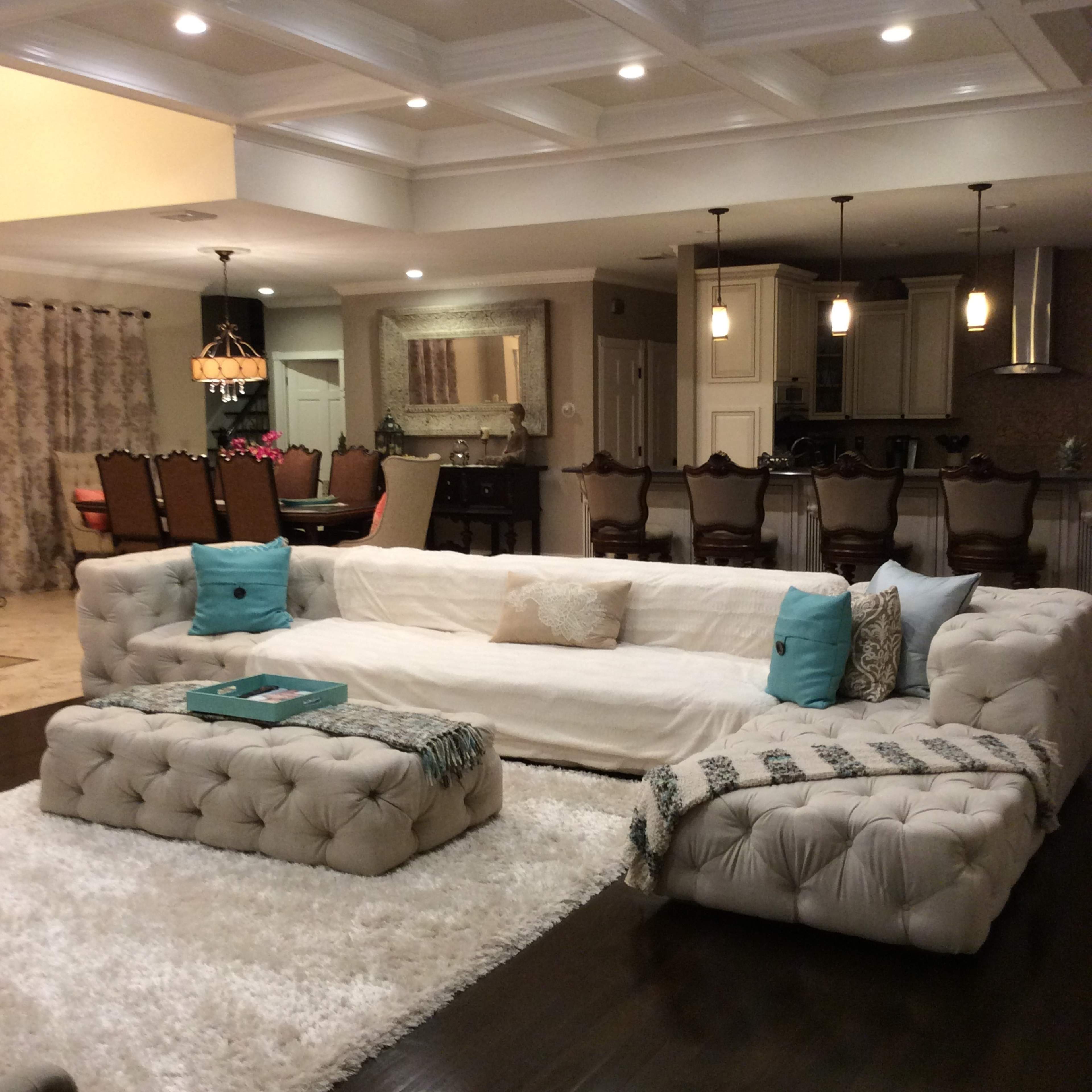 The living room features a large tufted sectional sofa, a plush white area rug, and an open kitchen area with barstools in the background.