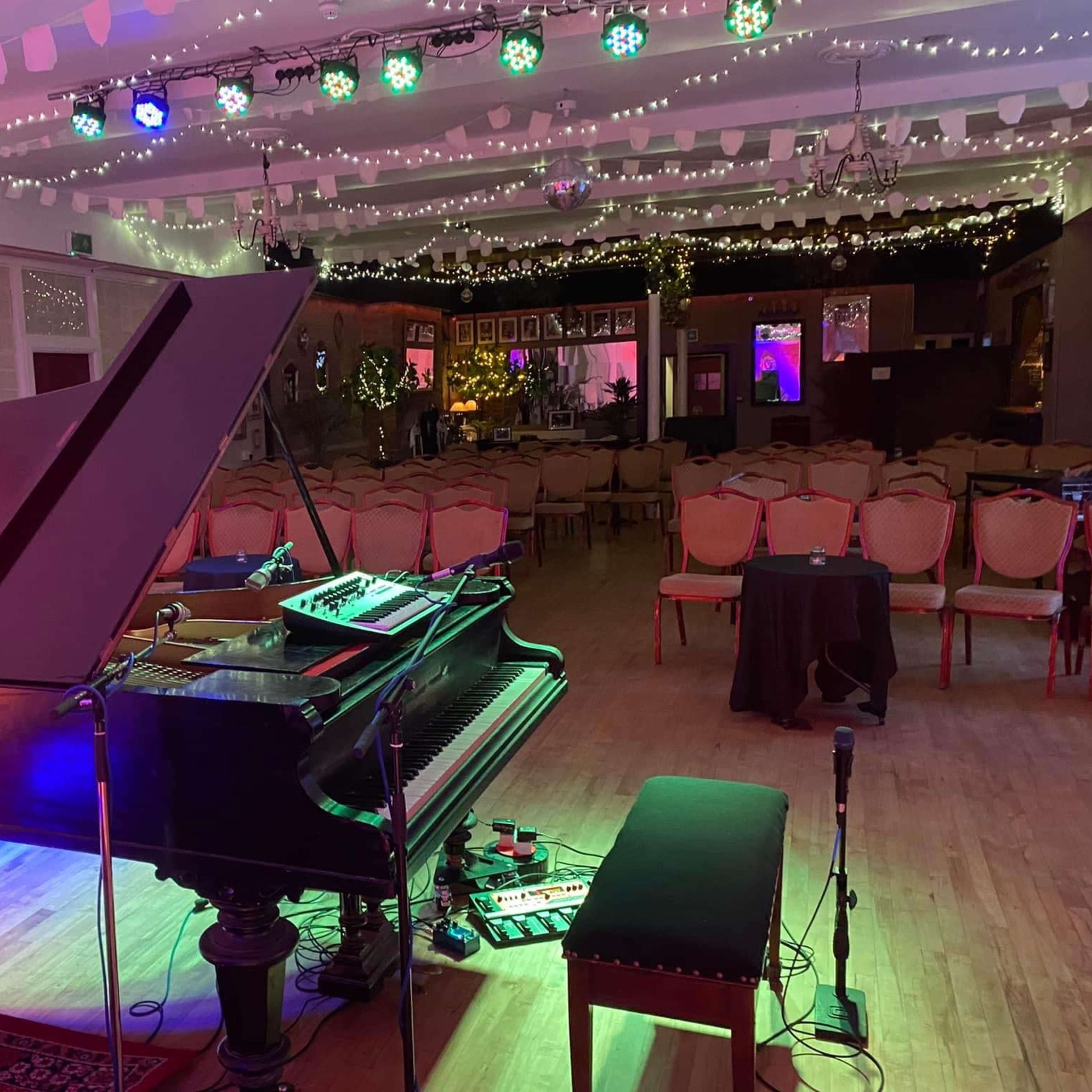 A grand piano is positioned in a well-lit room set up for an event, with chairs arranged in rows and decorative lights hanging from the ceiling.
