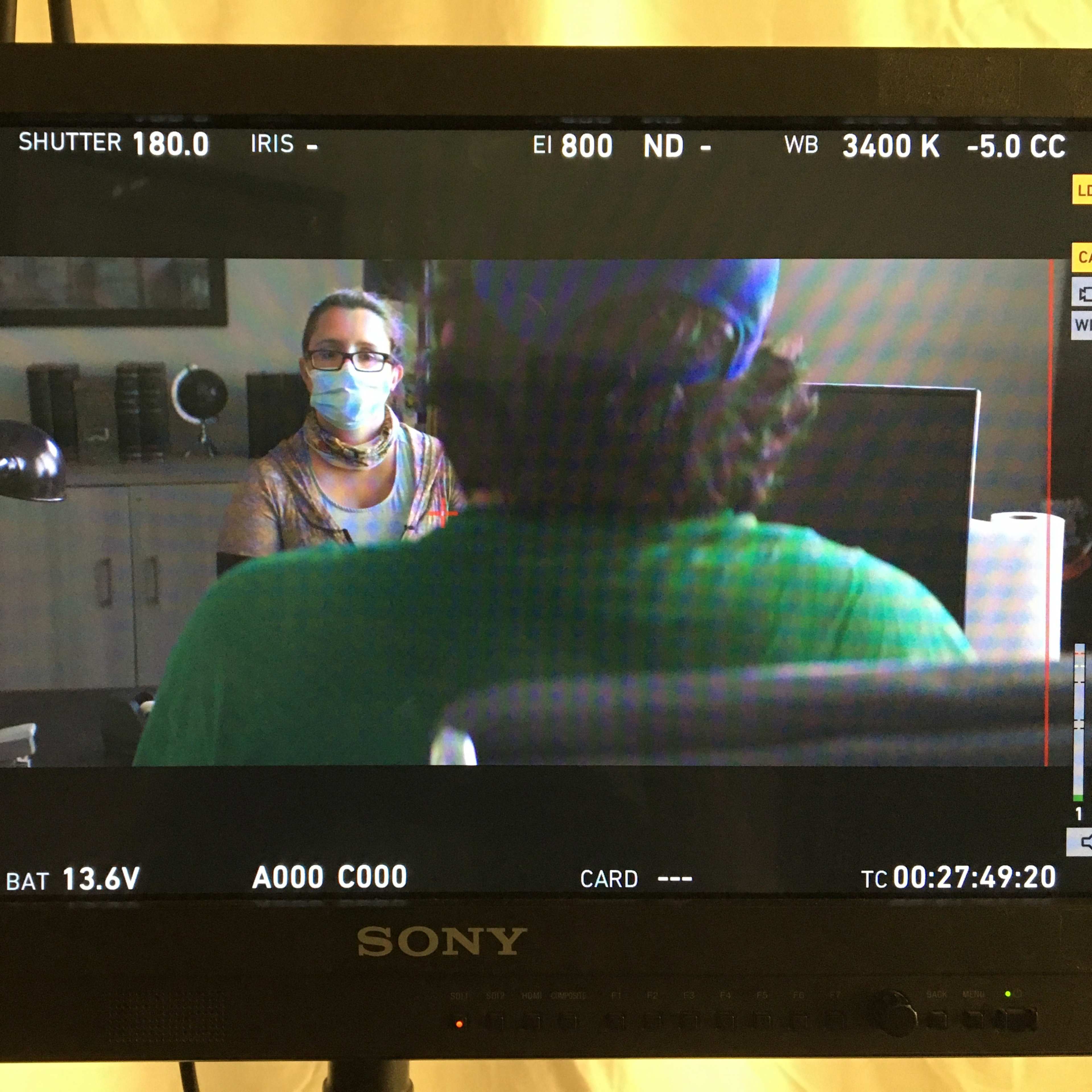 A camera monitor displaying a film scene with a woman wearing a mask talking to a person whose back is facing the camera.