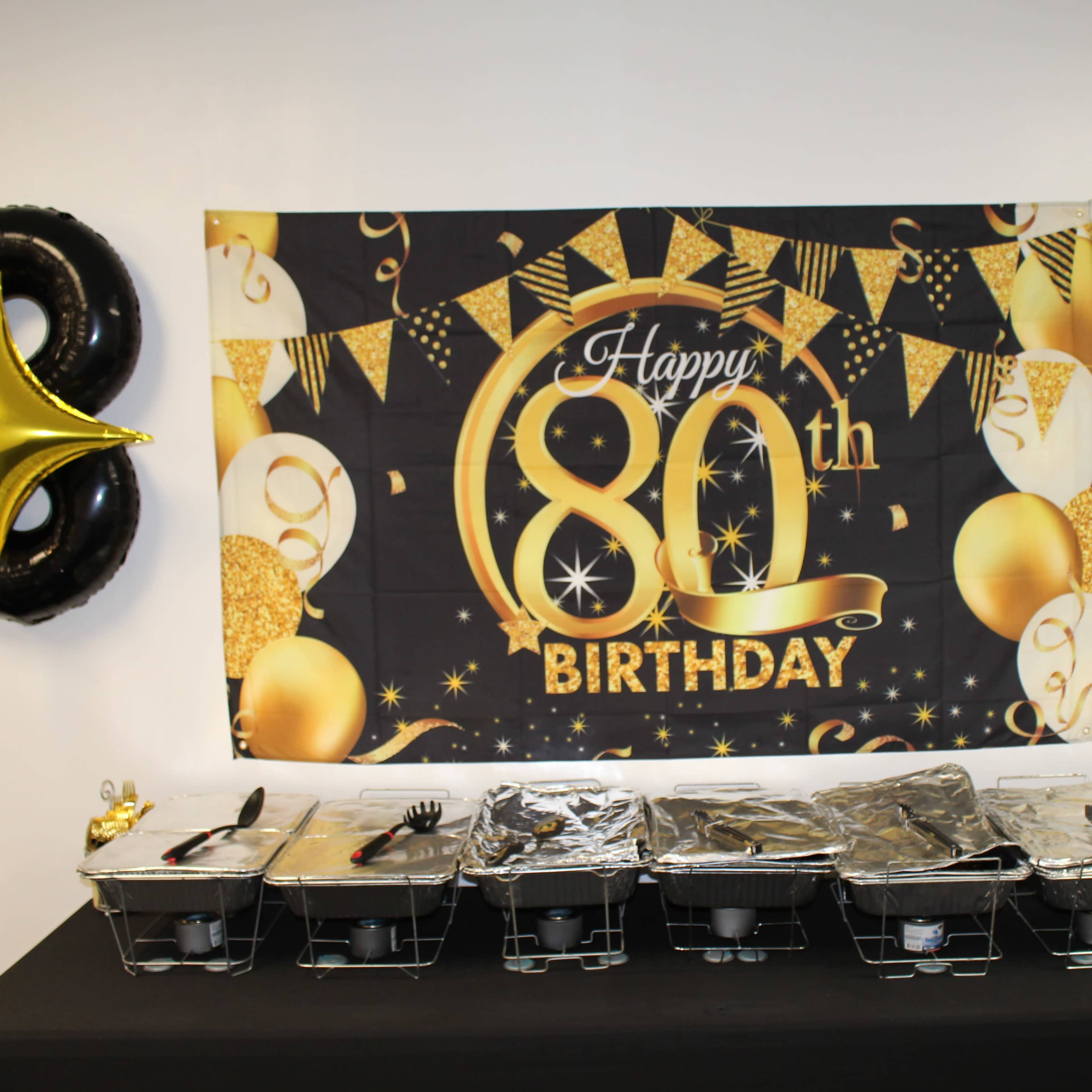 A table is set up for a birthday celebration featuring a large banner that reads "Happy 80th Birthday" along with gold and black decorations, and several covered food trays.