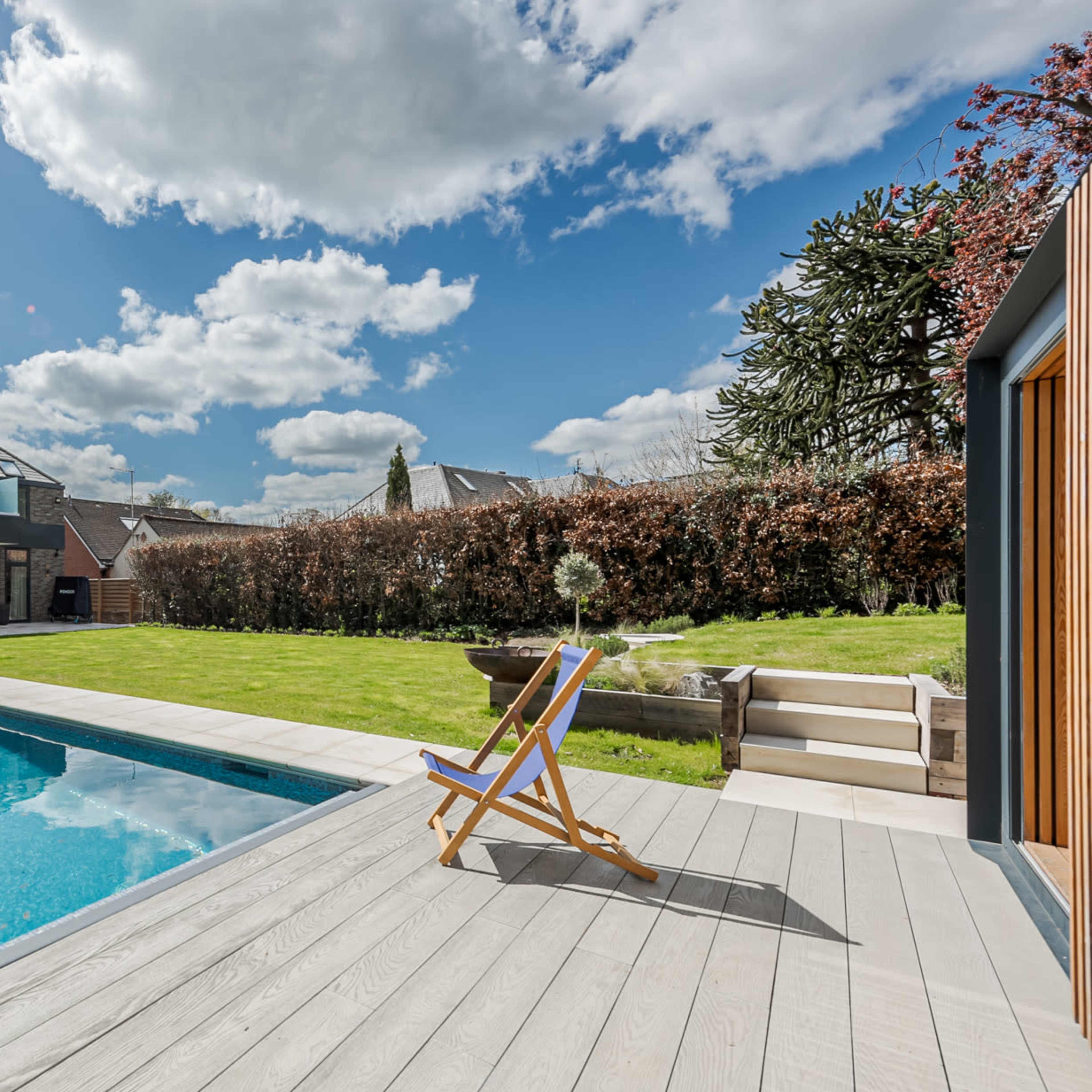 A modern house with large windows overlooks a rectangular swimming pool surrounded by a wooden deck and a manicured lawn.