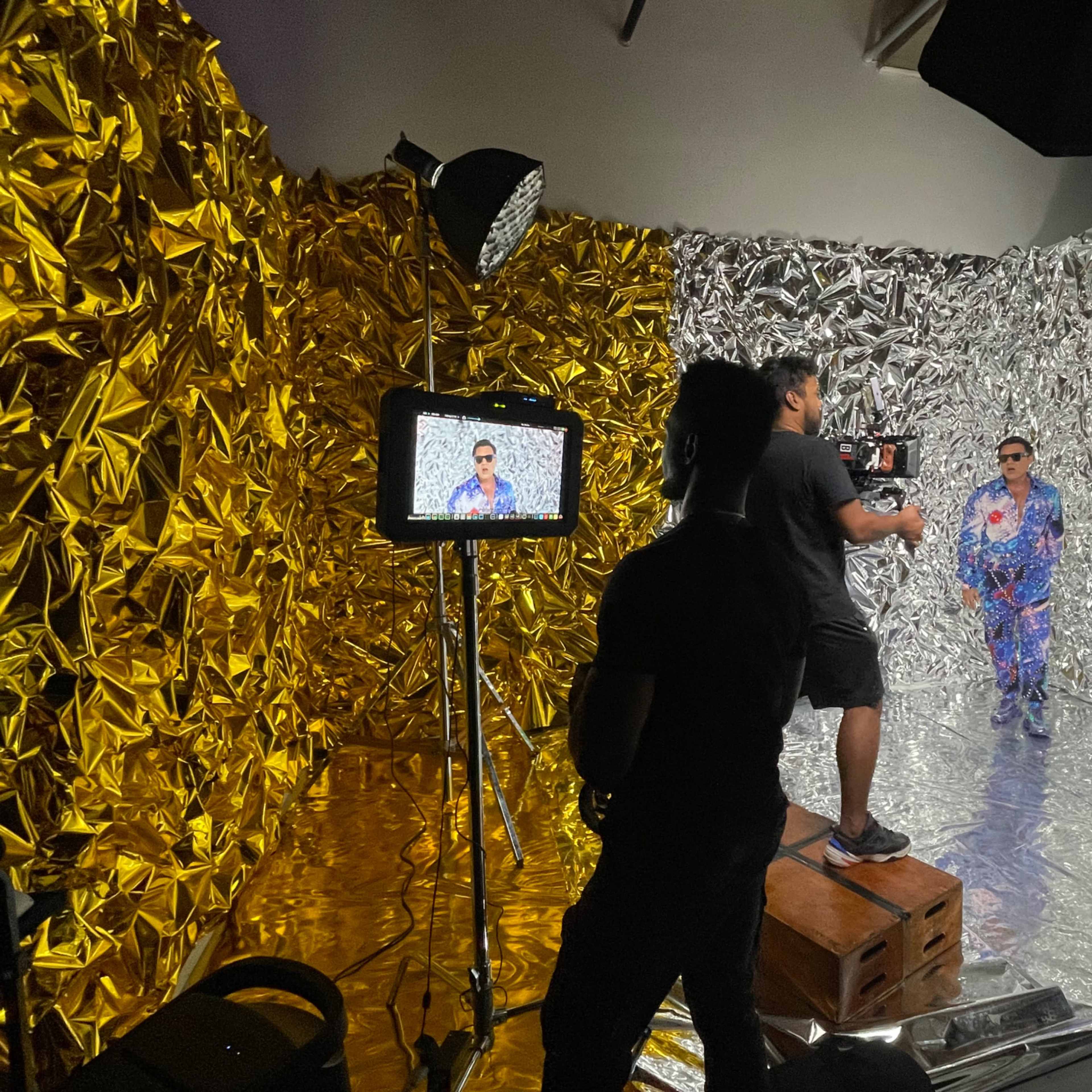 A man in a colorful outfit poses in a studio with metallic gold and silver backdrops while a crew member operates a camera.