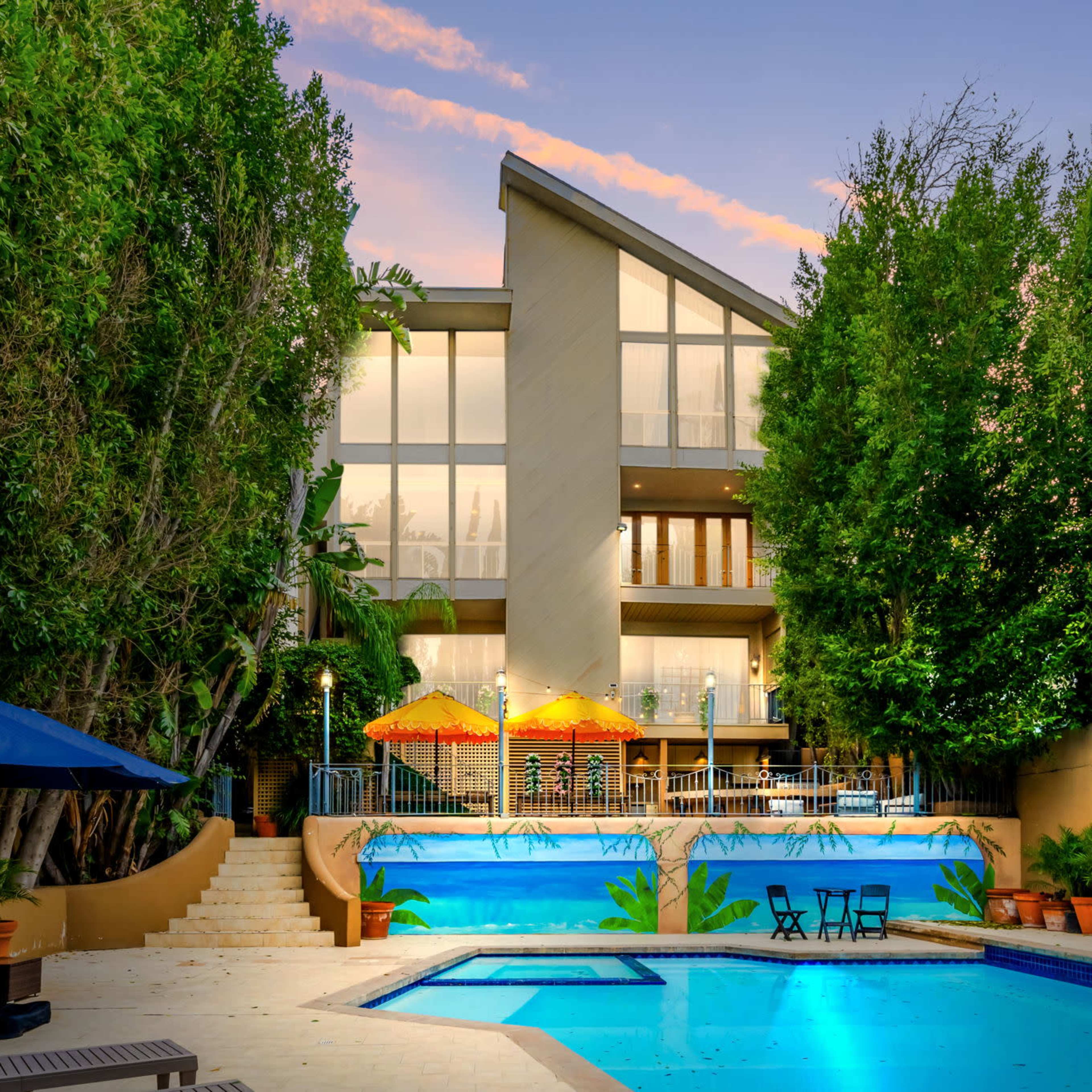 A modern house with large windows overlooks two swimming pools surrounded by greenery and lounge chairs.
