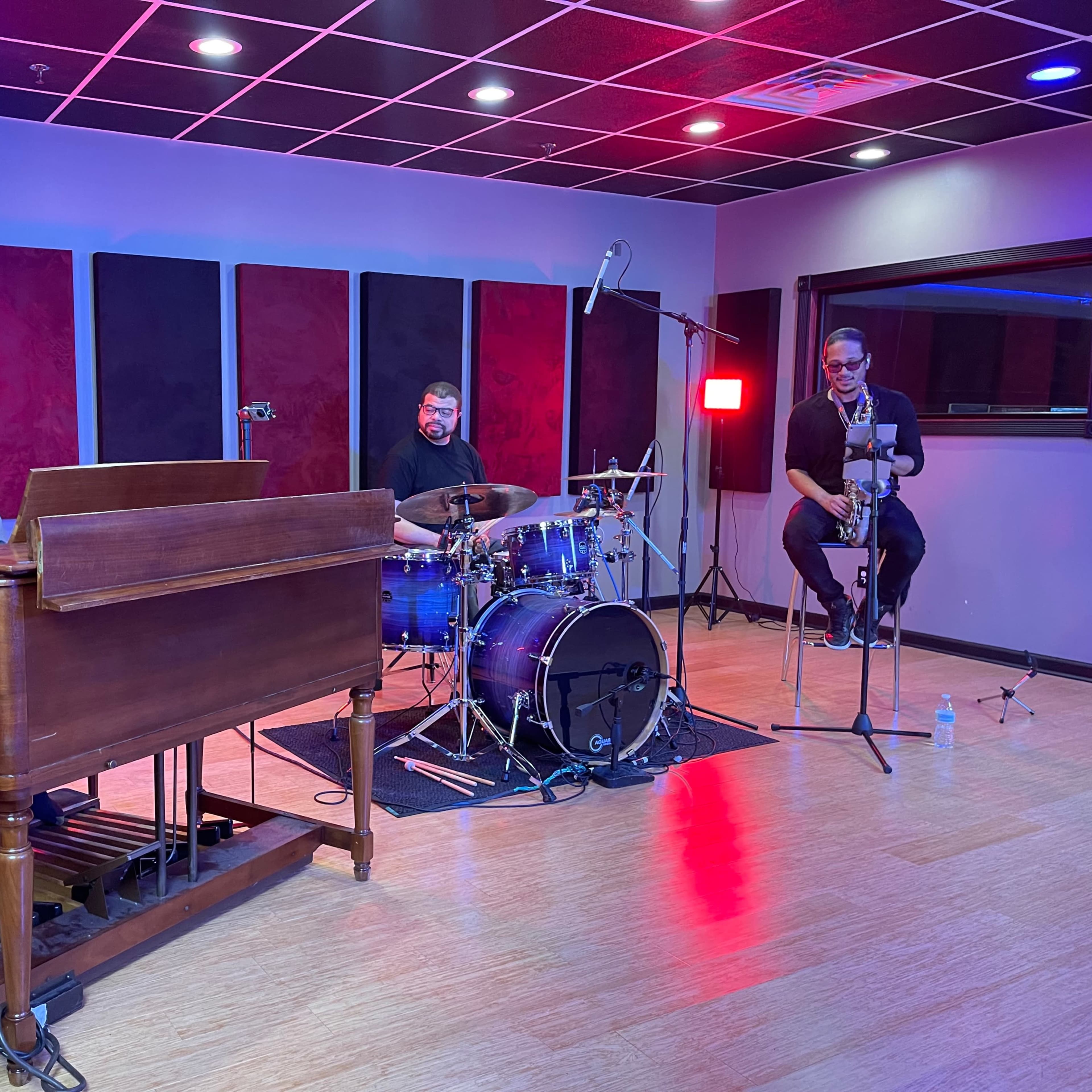 Three musicians perform in a recording studio, with a pianist on an organ, a drummer behind a drum set, and a saxophonist seated nearby.