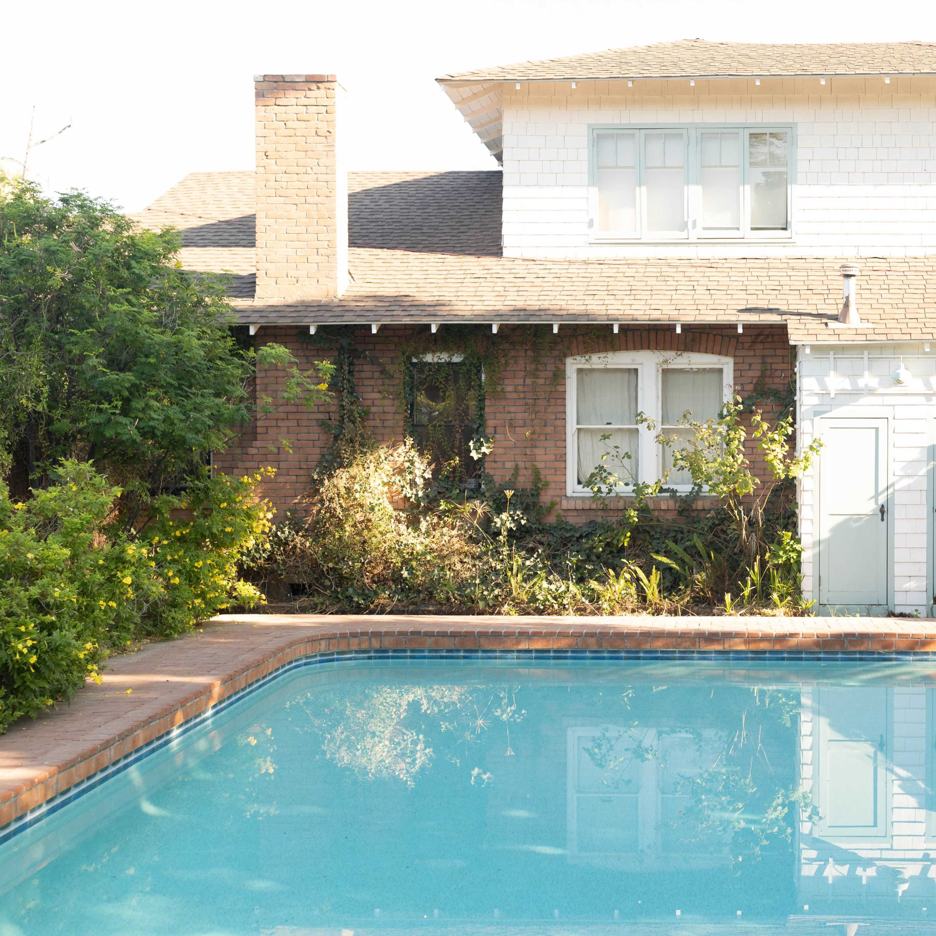 A clear swimming pool in front of a two-story house with a mix of brick and white siding, surrounded by lush greenery.