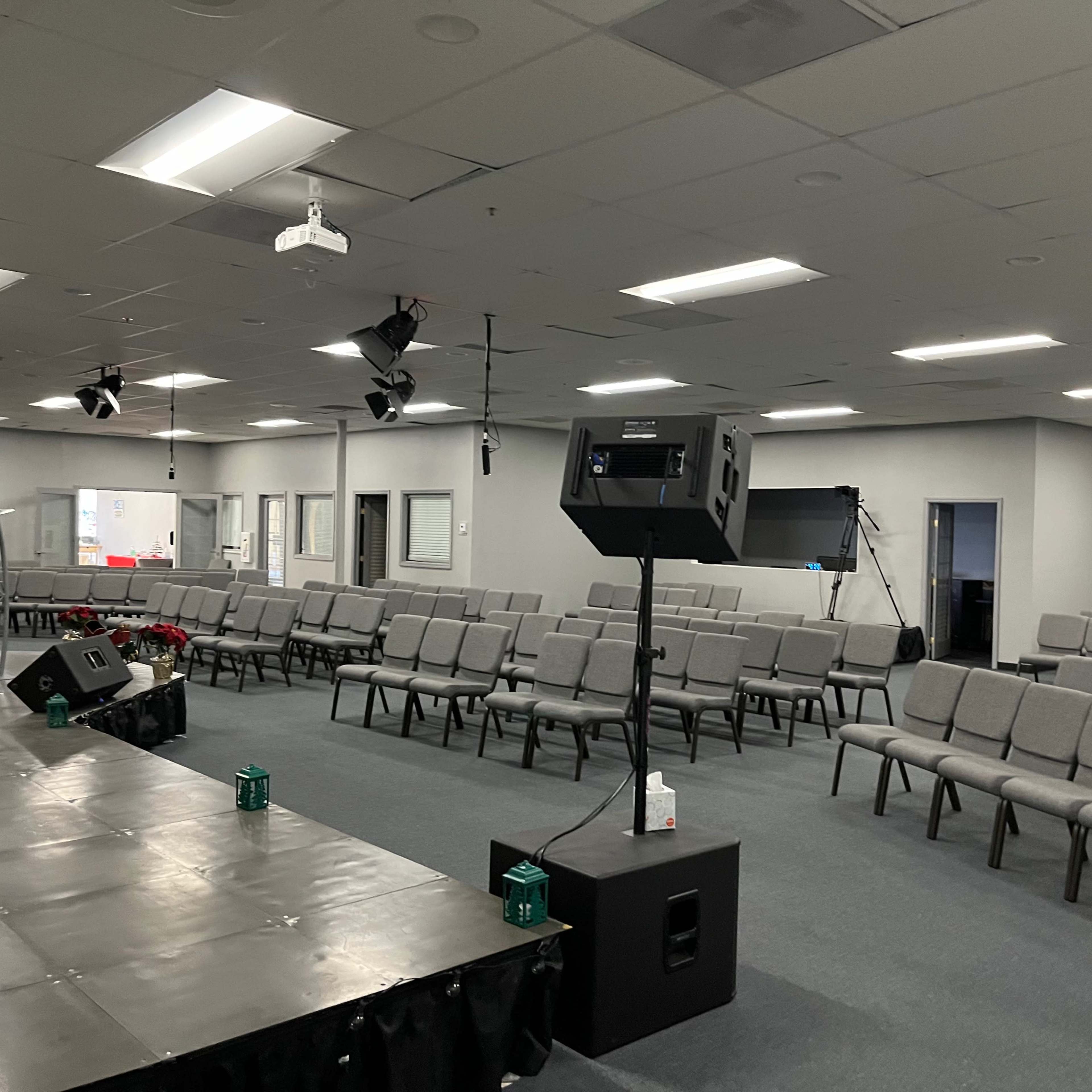 A spacious auditorium with rows of gray chairs facing a stage, and various lighting and audio equipment set up around the room.