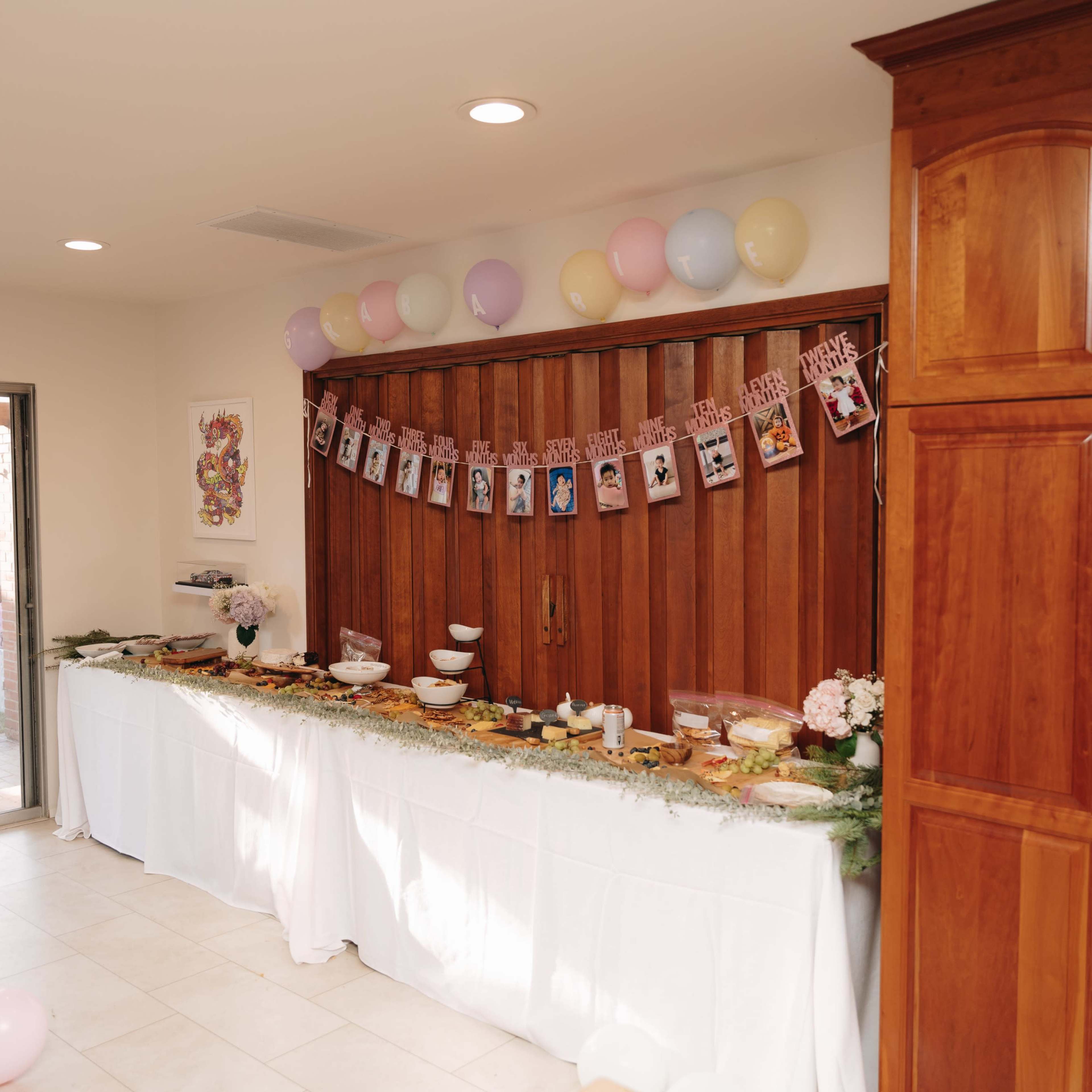 A festive table laden with food and decorated with photos and balloons is set against a wooden wall in a well-lit room.