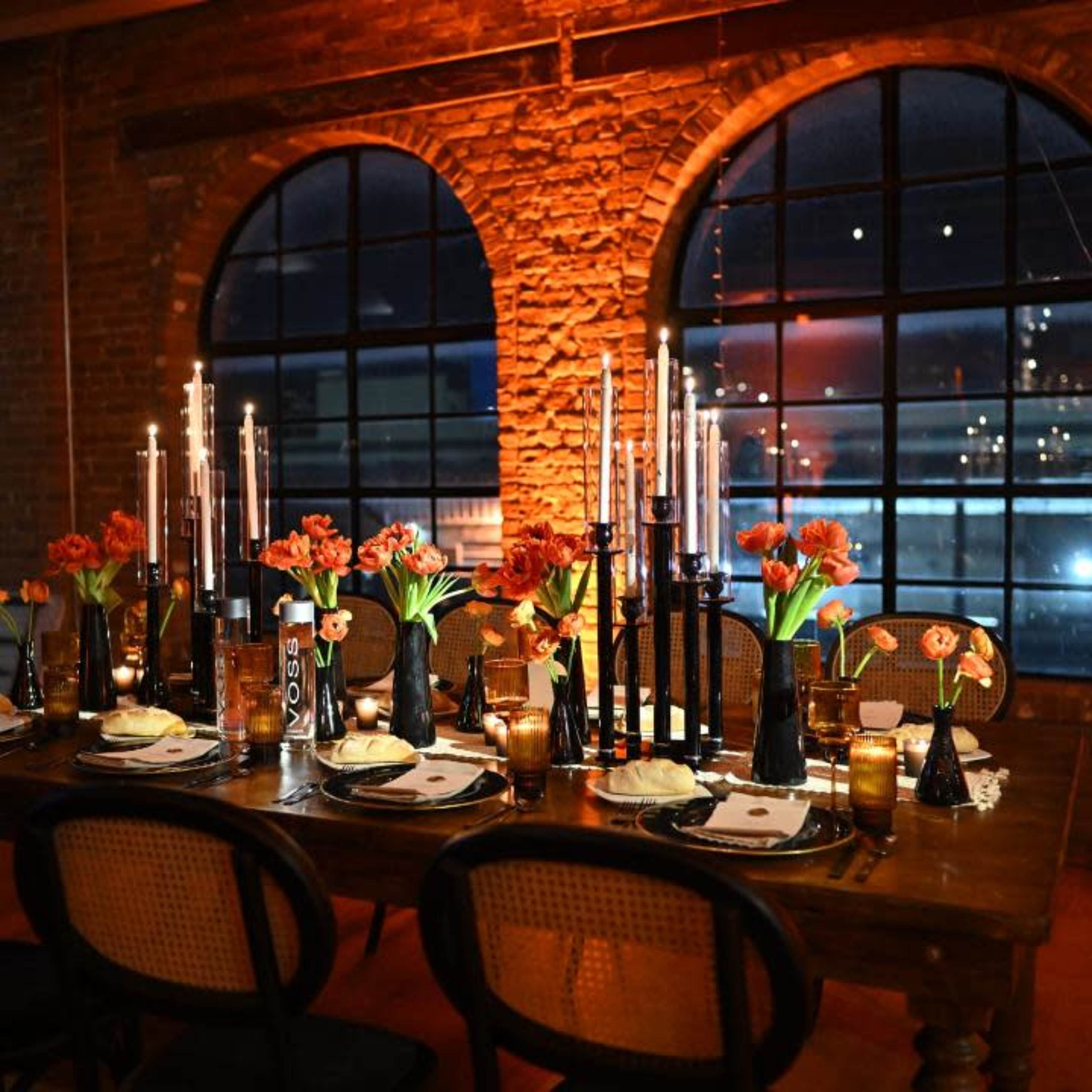 A long dining table is set with floral centerpieces, candles, and tableware in a brick-walled room with large arched windows.