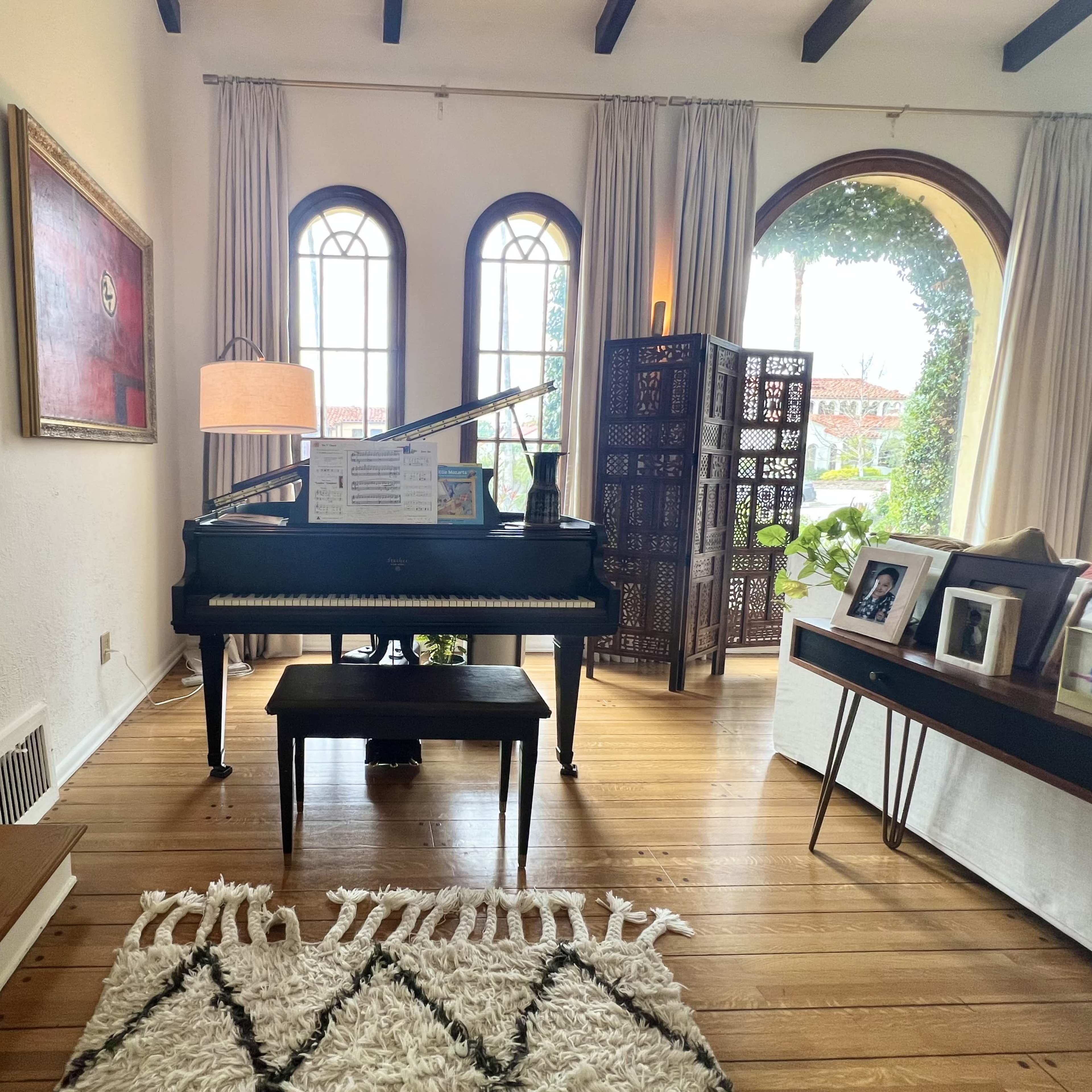A bright living room featuring a piano with sheet music on it, surrounded by windows and a decorative folding screen.