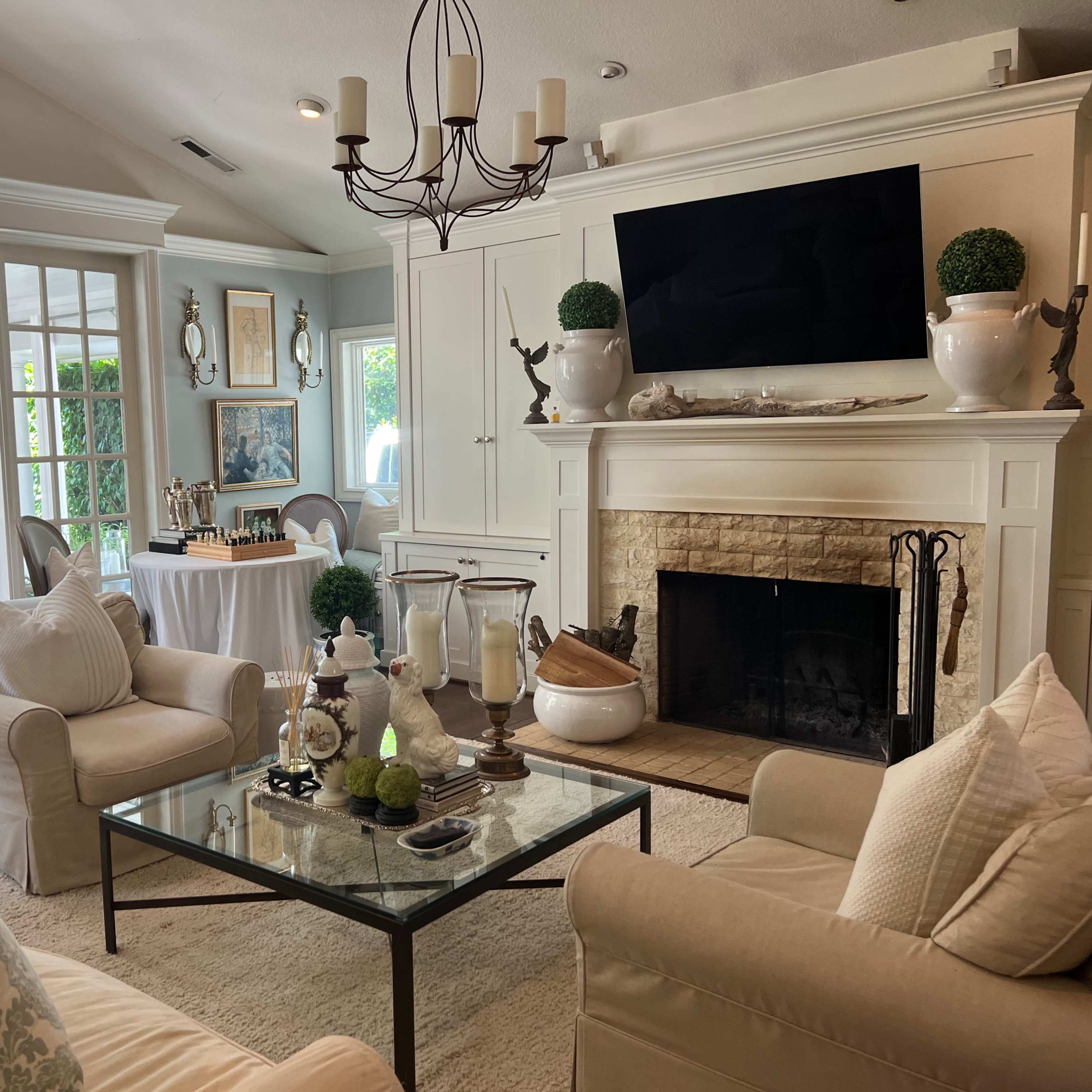 A light-filled living room with a cream-colored sofa, a glass coffee table, and a stone fireplace adorned with decorative elements, leading to a sunlit sunroom through French doors.