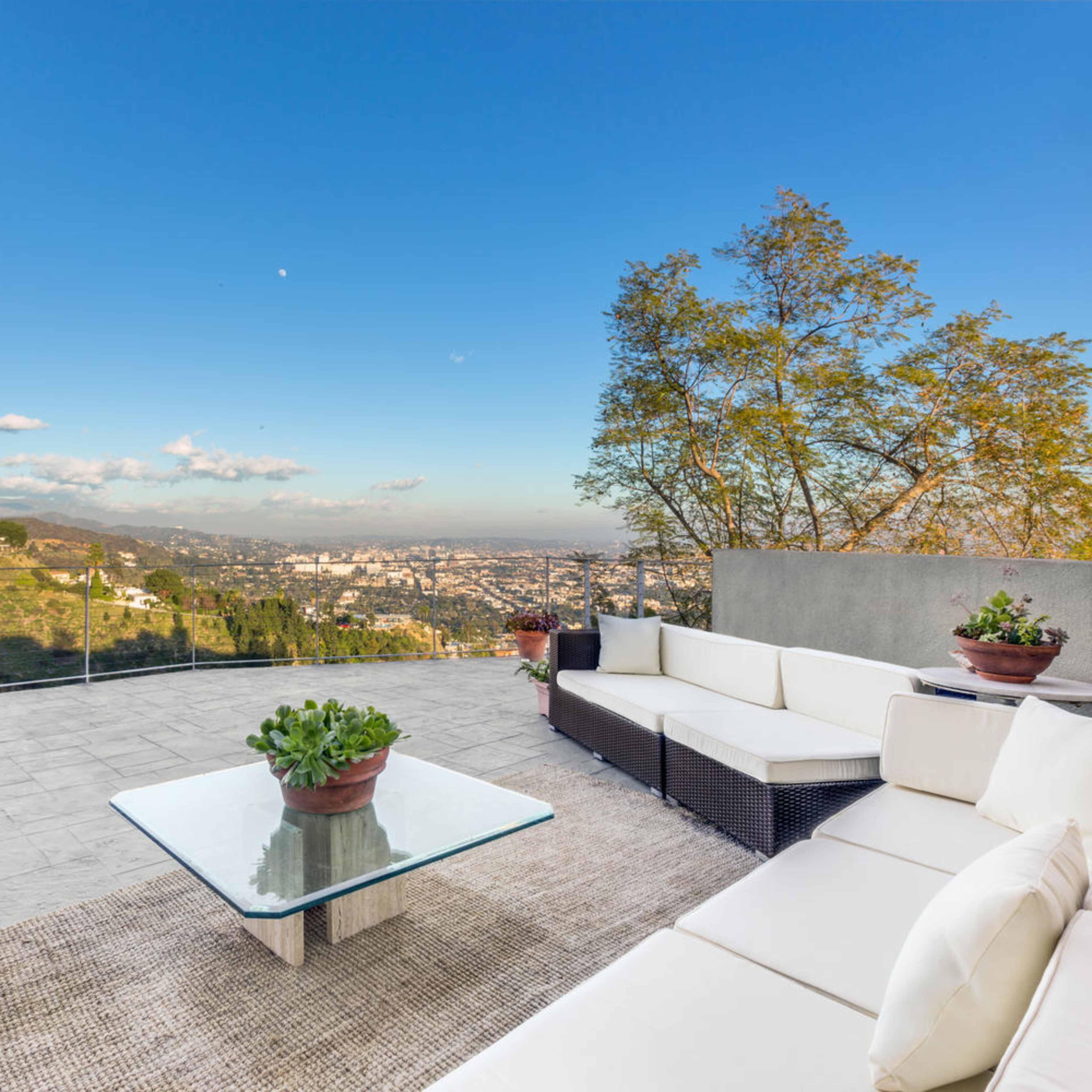 A modern outdoor patio with a glass table and white sofas, overlooking a city view and mountains in the background.