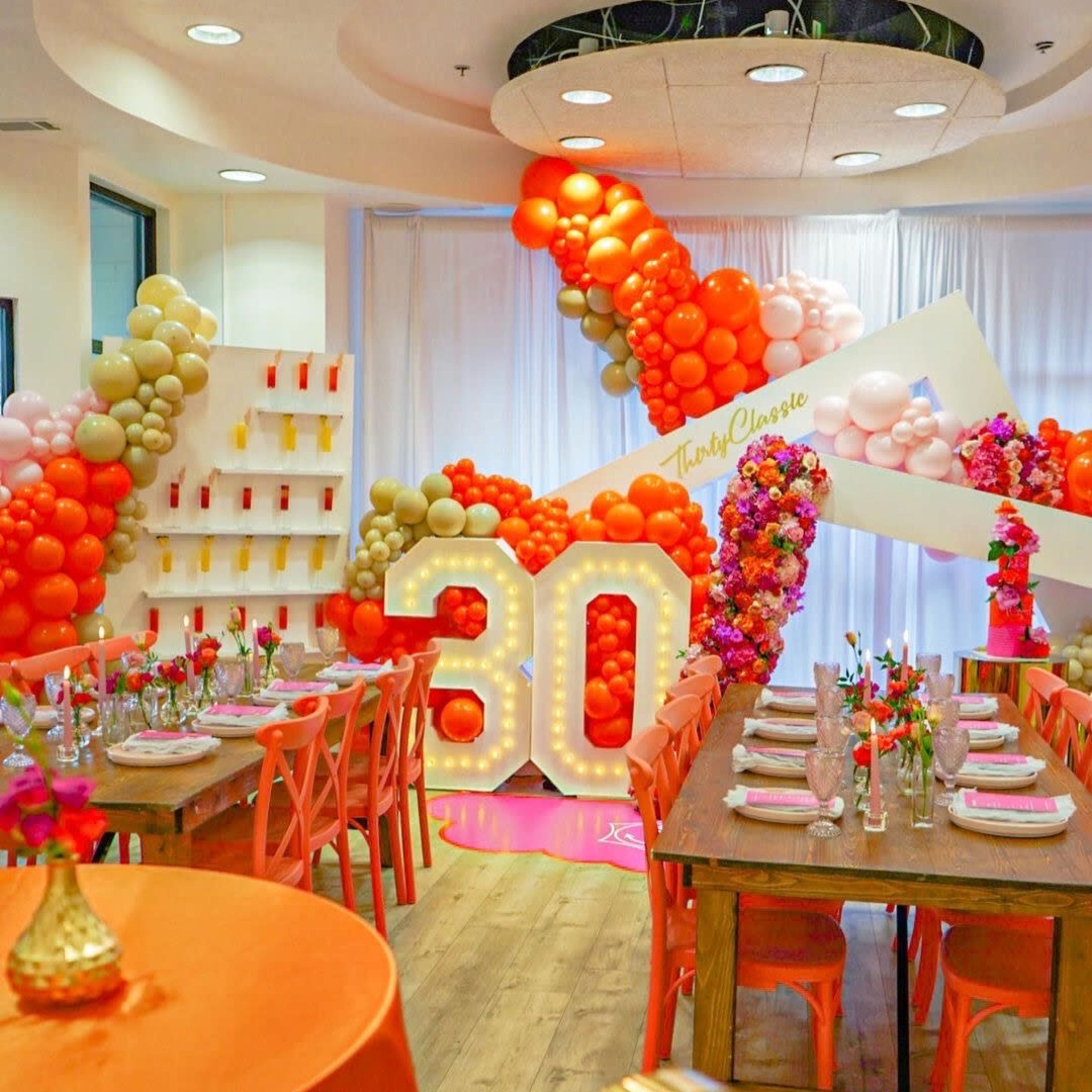 A decorated event space features two large tables with orange tablecloths, surrounded by colorful balloon arrangements and a prominent "30" display.