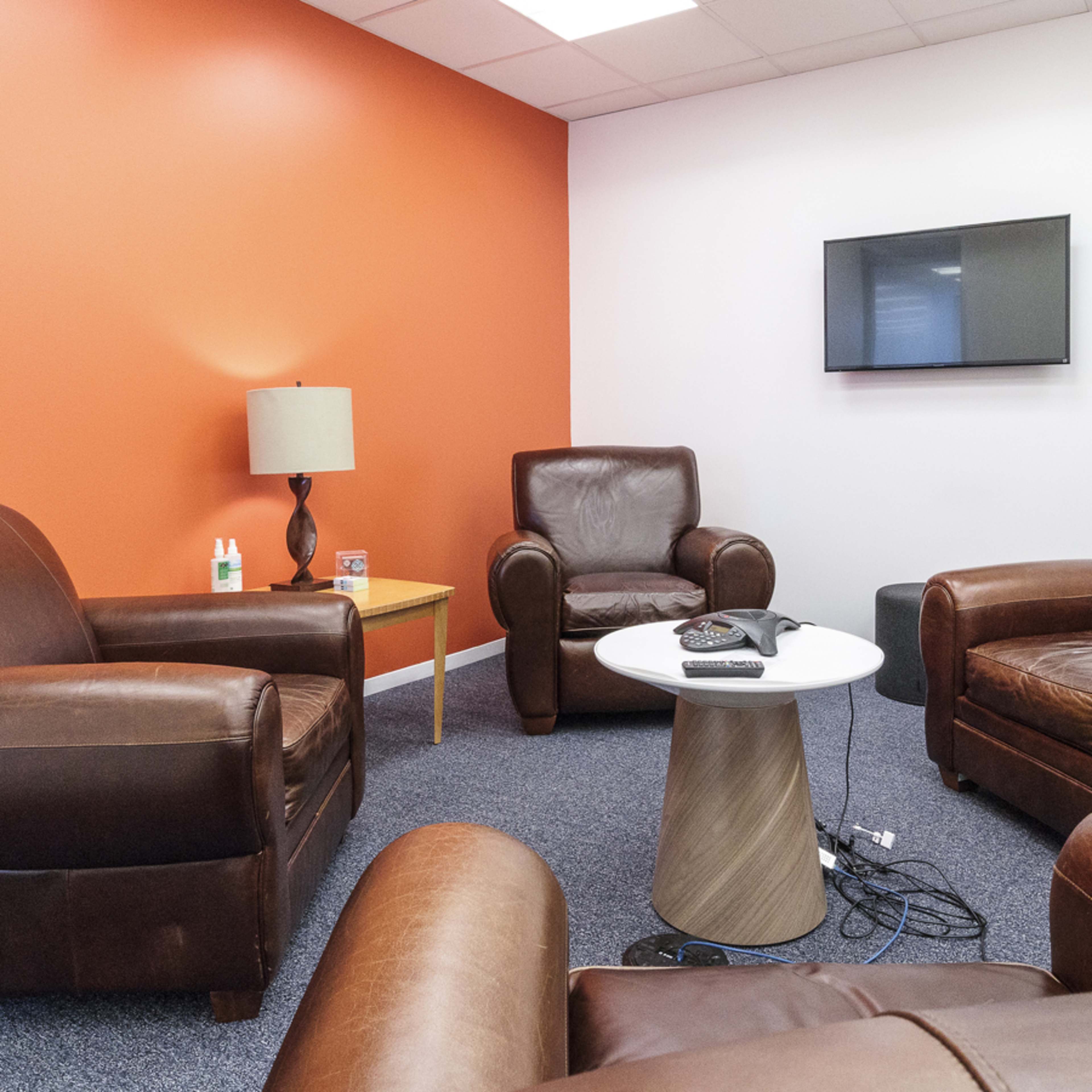 A small lounge area with two leather couches, a coffee table, a lamp, and a wall-mounted television, all set against an orange wall.
