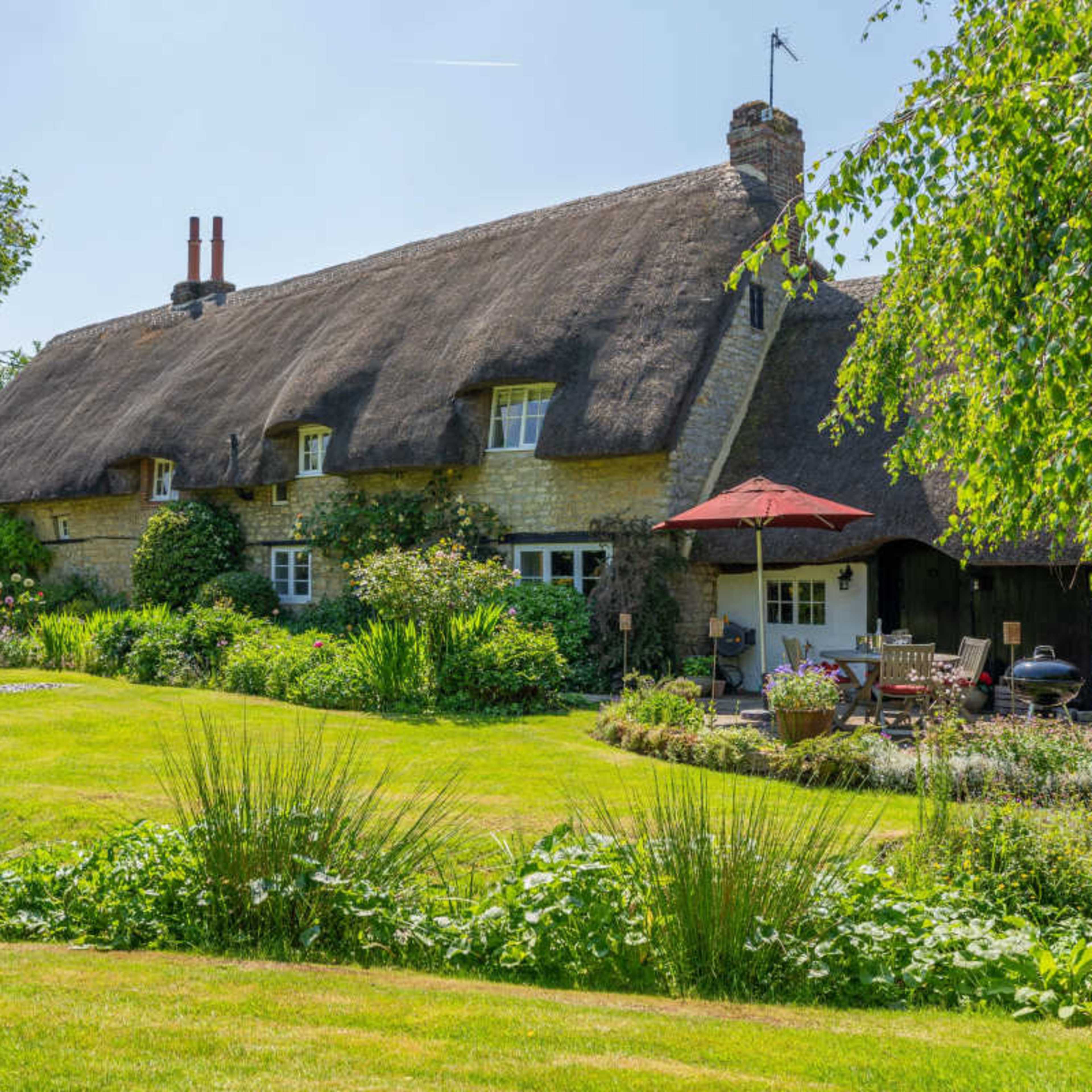 A thatched-roof cottage is surrounded by a lush garden featuring various plants and a red umbrella over a seating area.