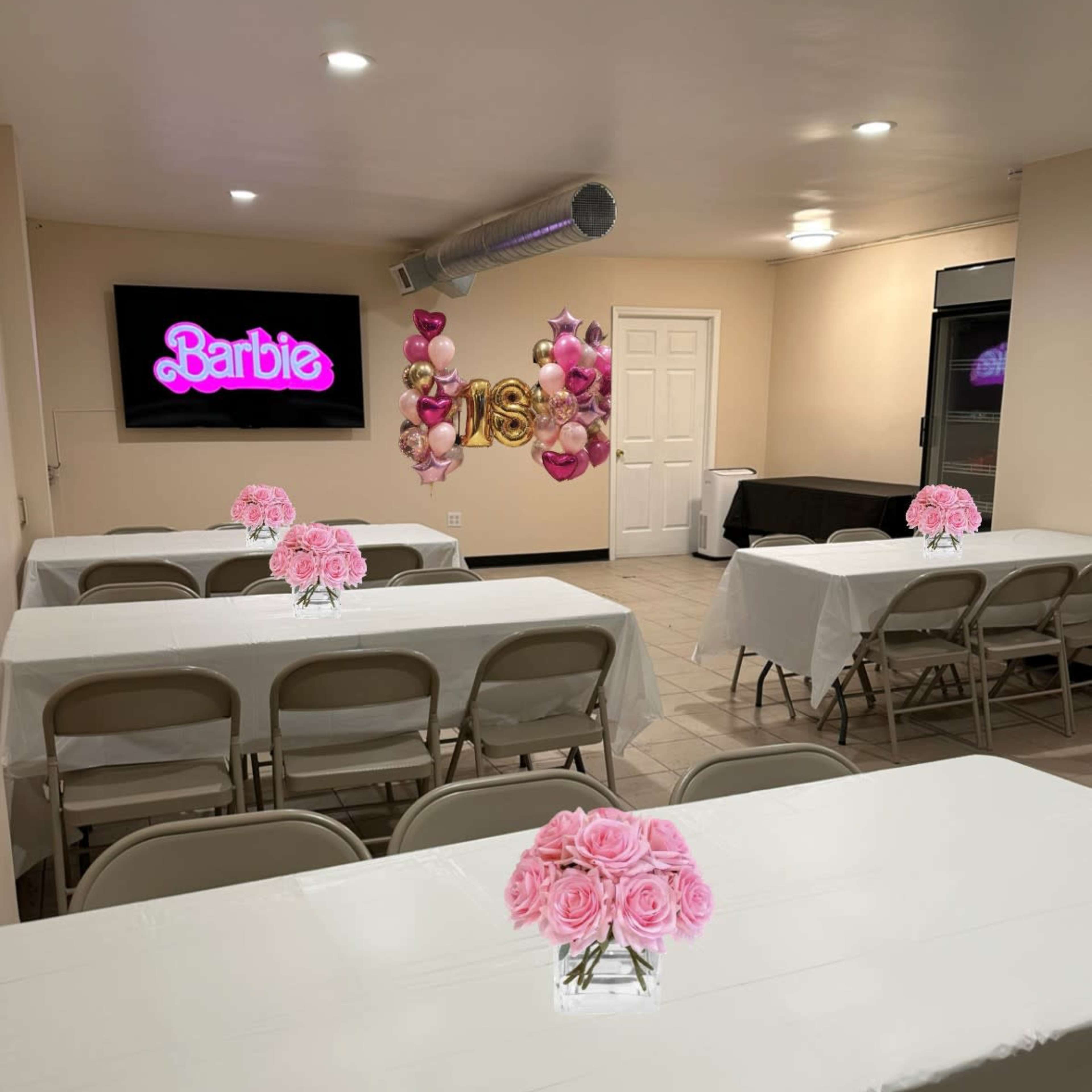 A decorated room set up for a celebration, featuring white tables with pink rose centerpieces and a large "Barbie" sign on the wall.