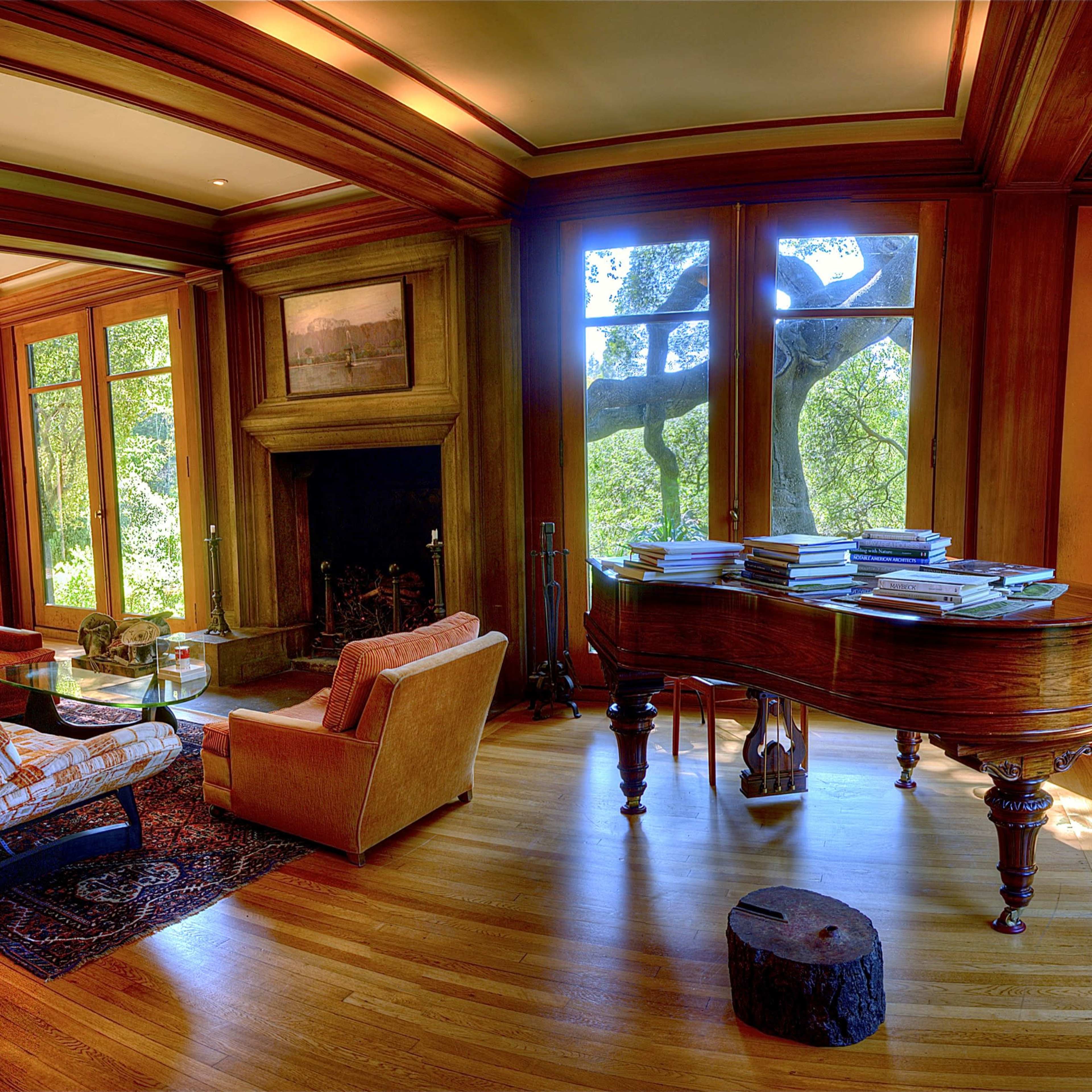 A spacious living room featuring large windows that provide a view of a garden, a grand piano, and seating areas with a wooden floor.
