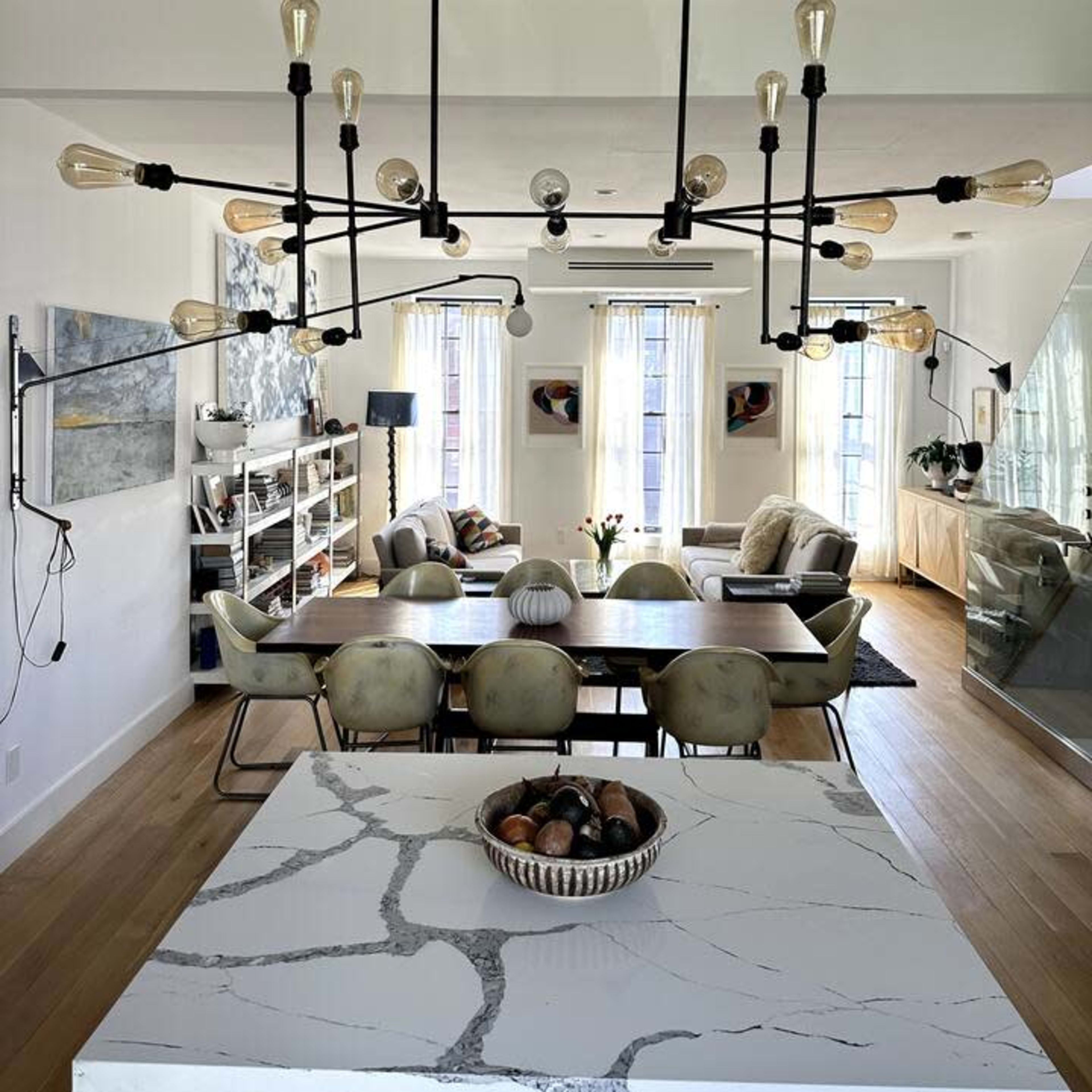 A modern dining area with a large table, surrounded by chairs, and an industrial-style chandelier hanging above, with a kitchen island featuring a marble top in the foreground.