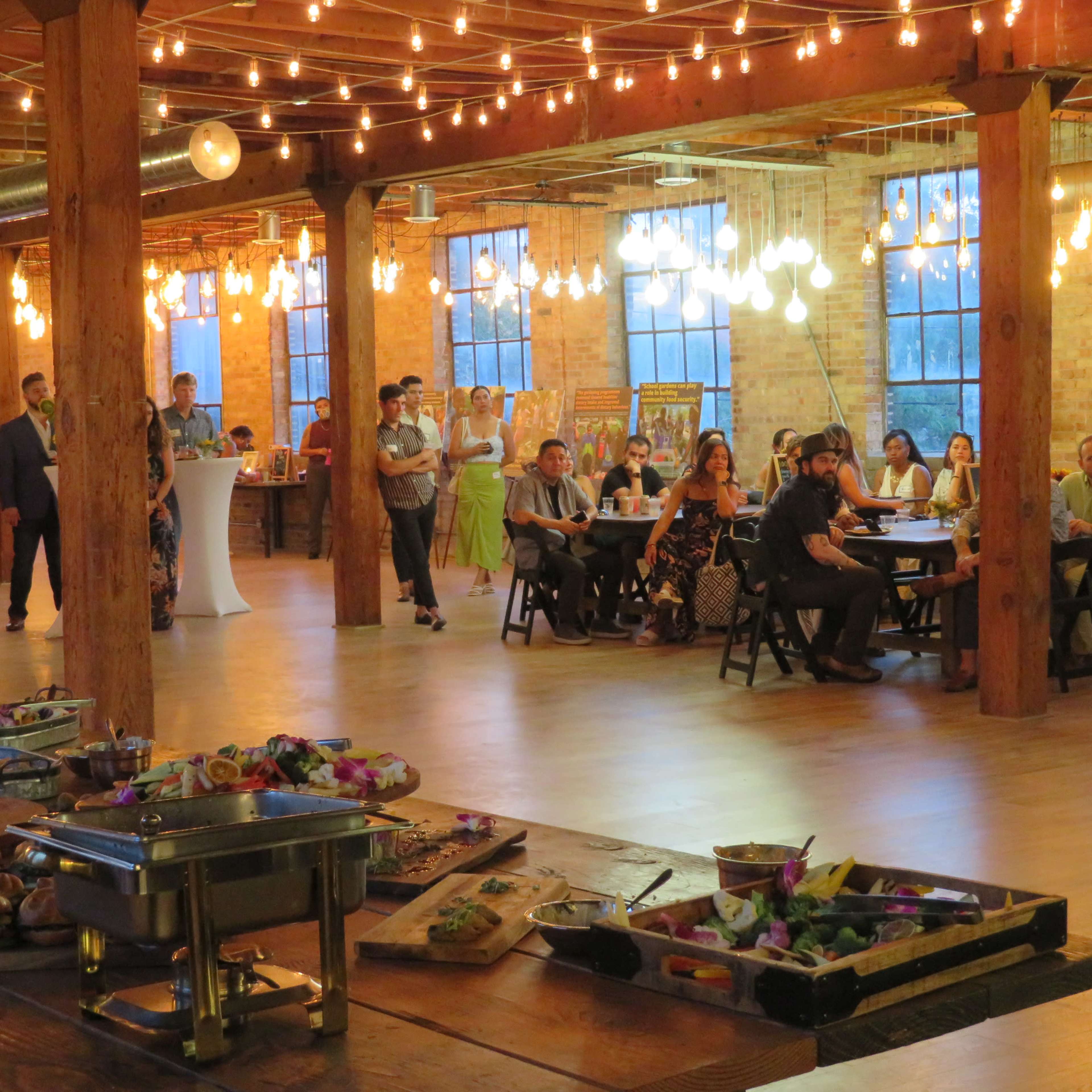 A spacious event venue features guests mingling, seated at tables, and a buffet with an array of food prominently displayed in the foreground.