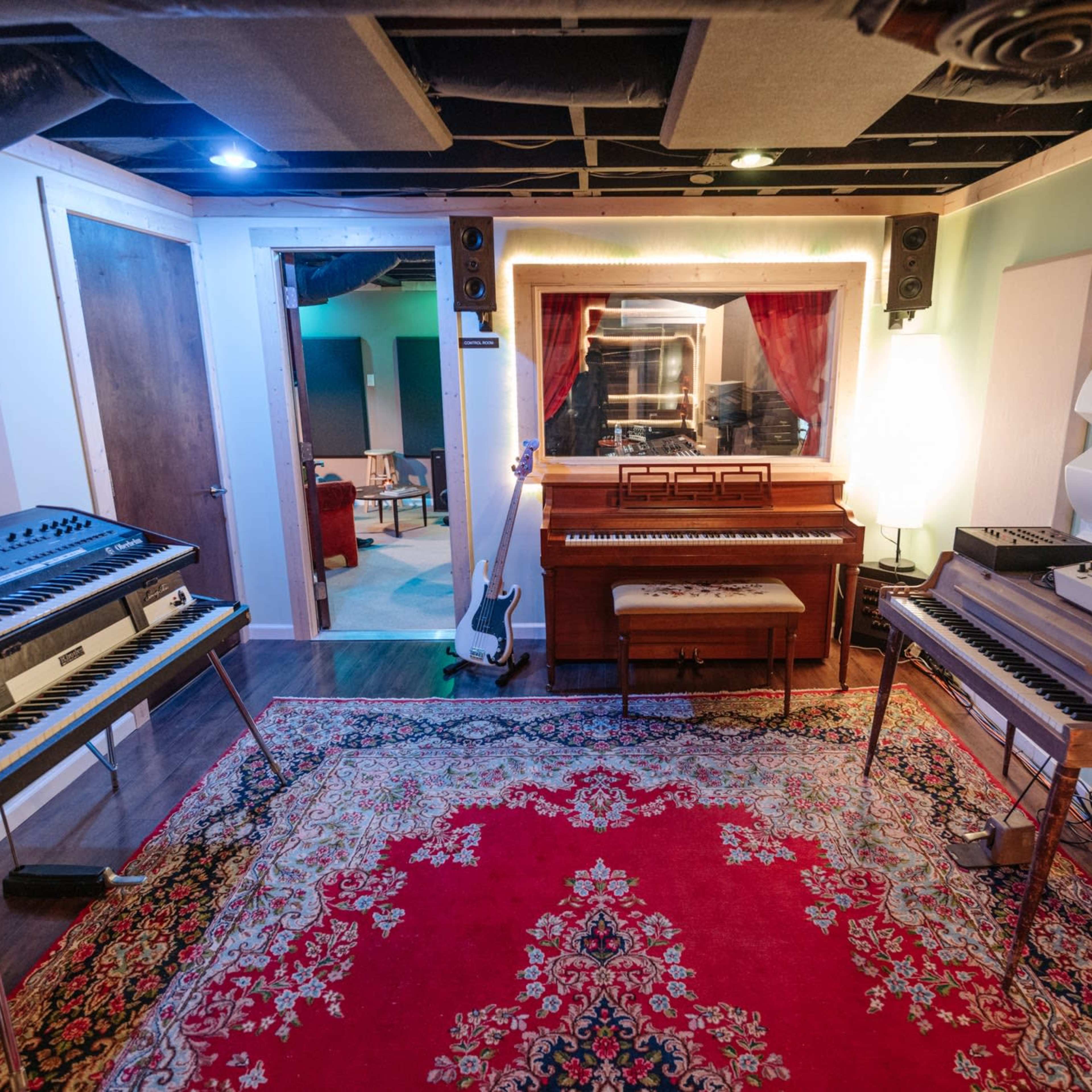 A music studio with synthesizers and a piano arranged around a central area featuring a large red area rug.