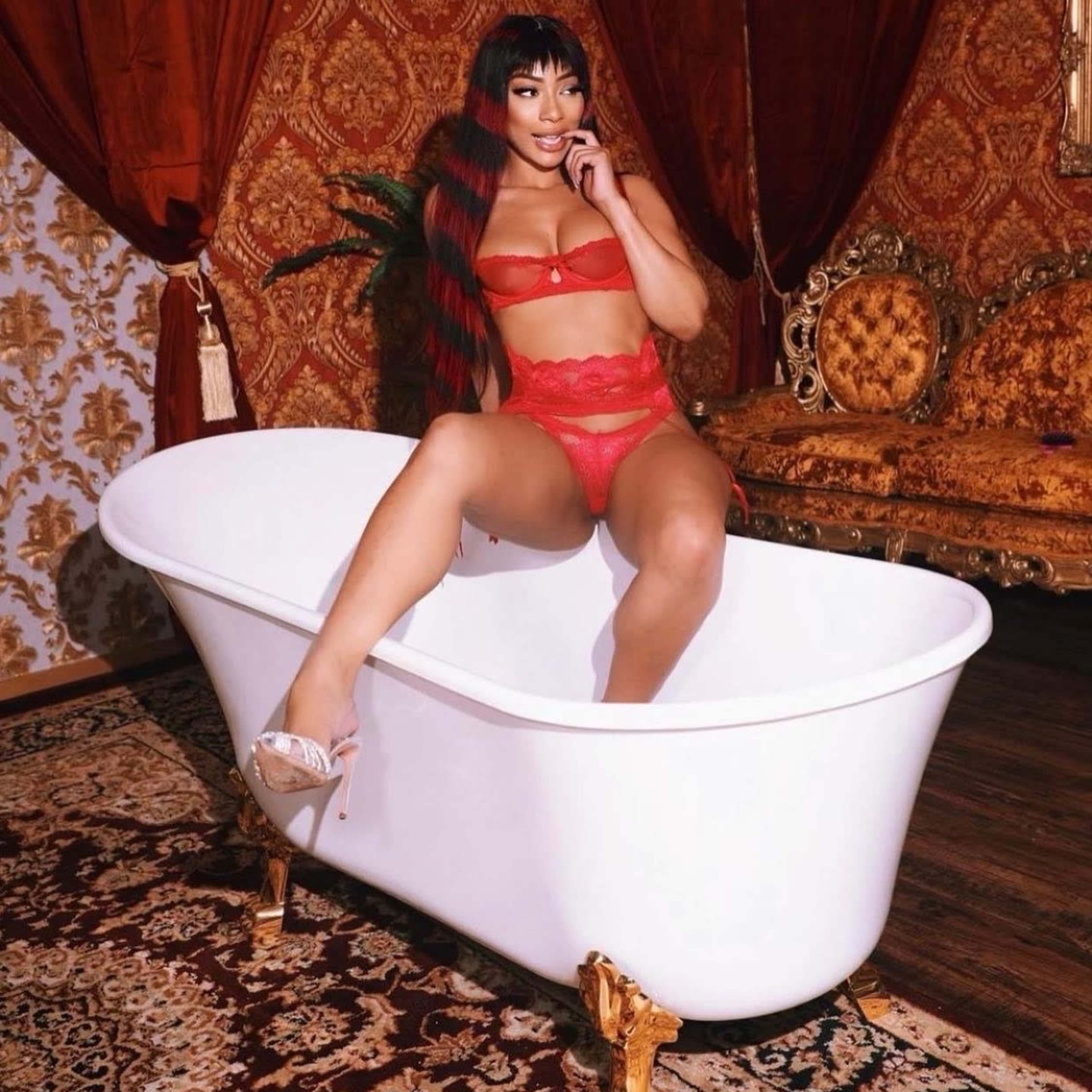 A woman in red lingerie poses playfully in a white freestanding bathtub against a richly decorated backdrop with ornate furniture and patterned walls.