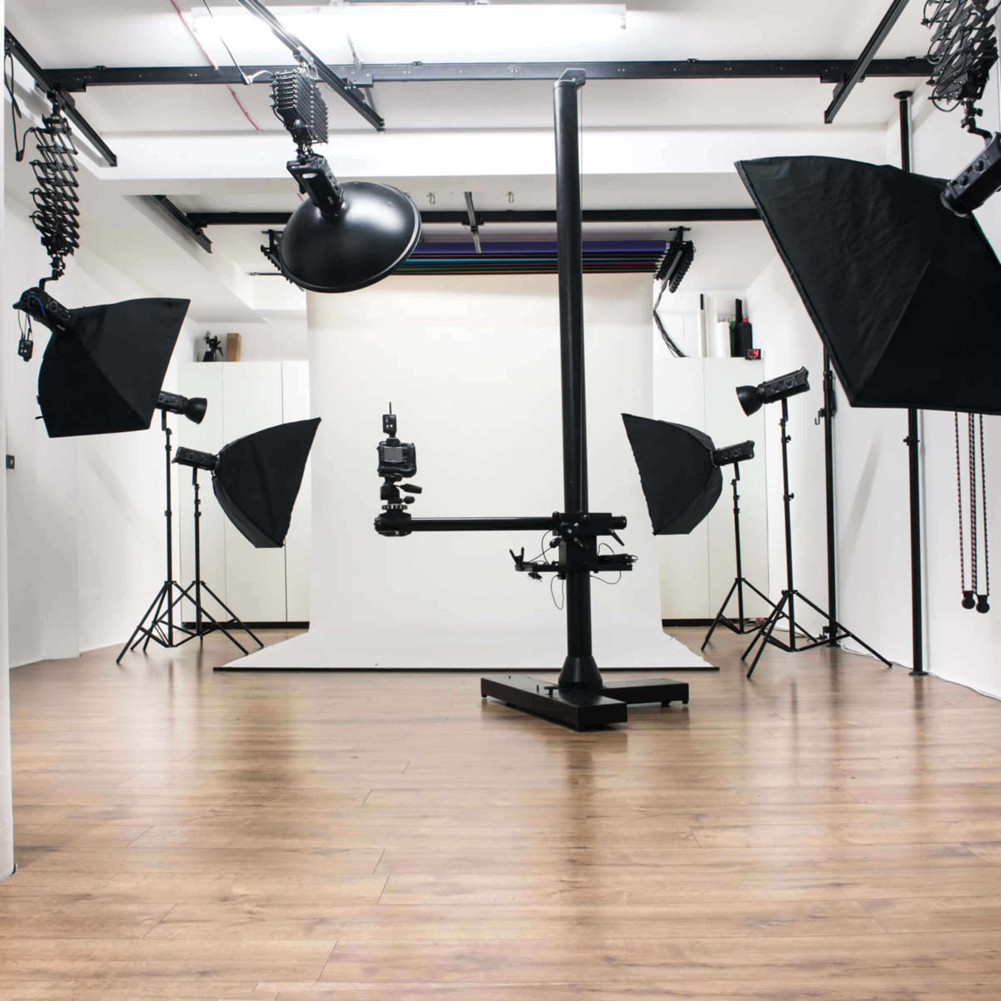 A photography studio features a backdrop setup with multiple light sources, a camera on a stand, and a workspace with a desk and chair.