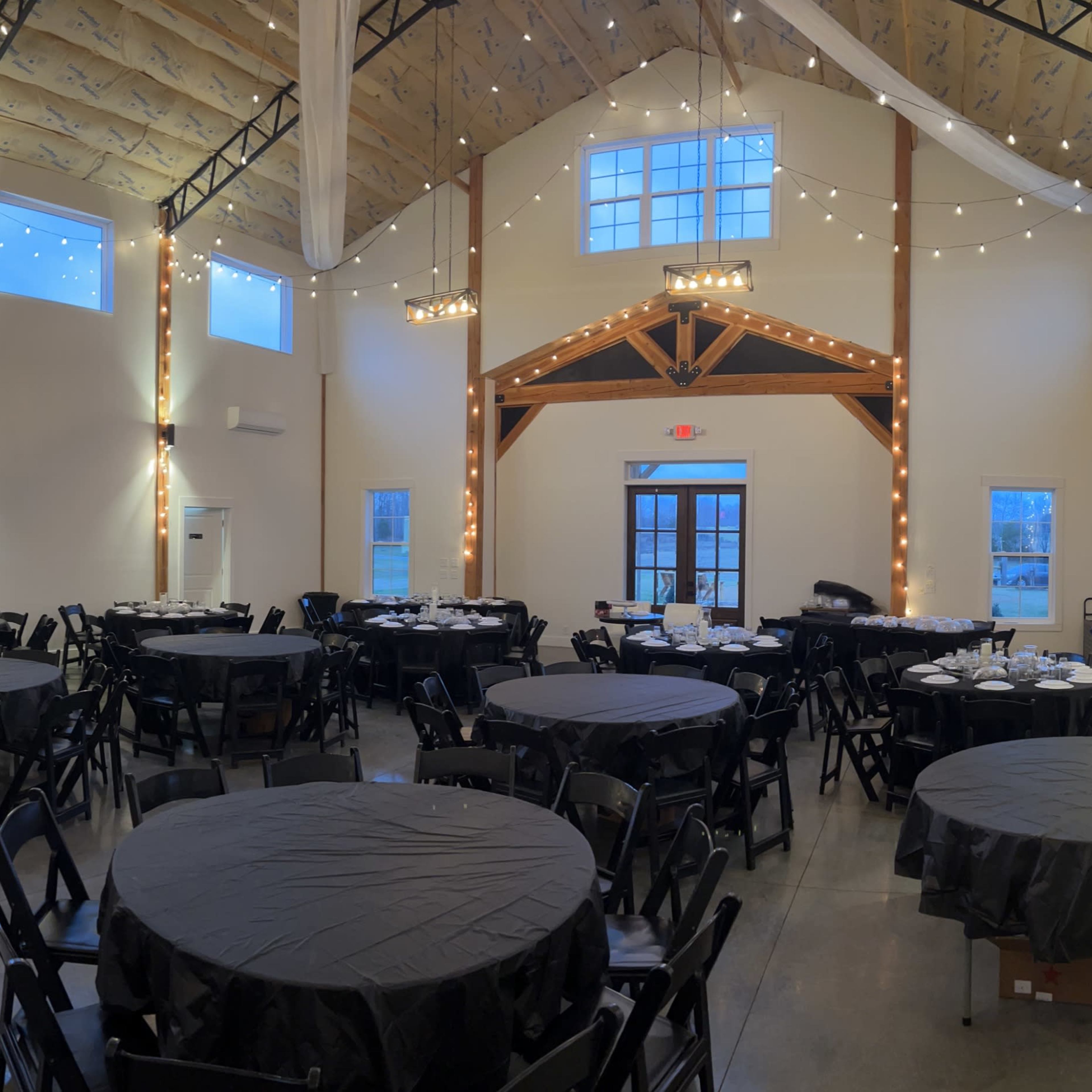 A spacious event hall with black round tables arranged throughout, string lights illuminating the high ceiling, and large windows allowing natural light to enter.