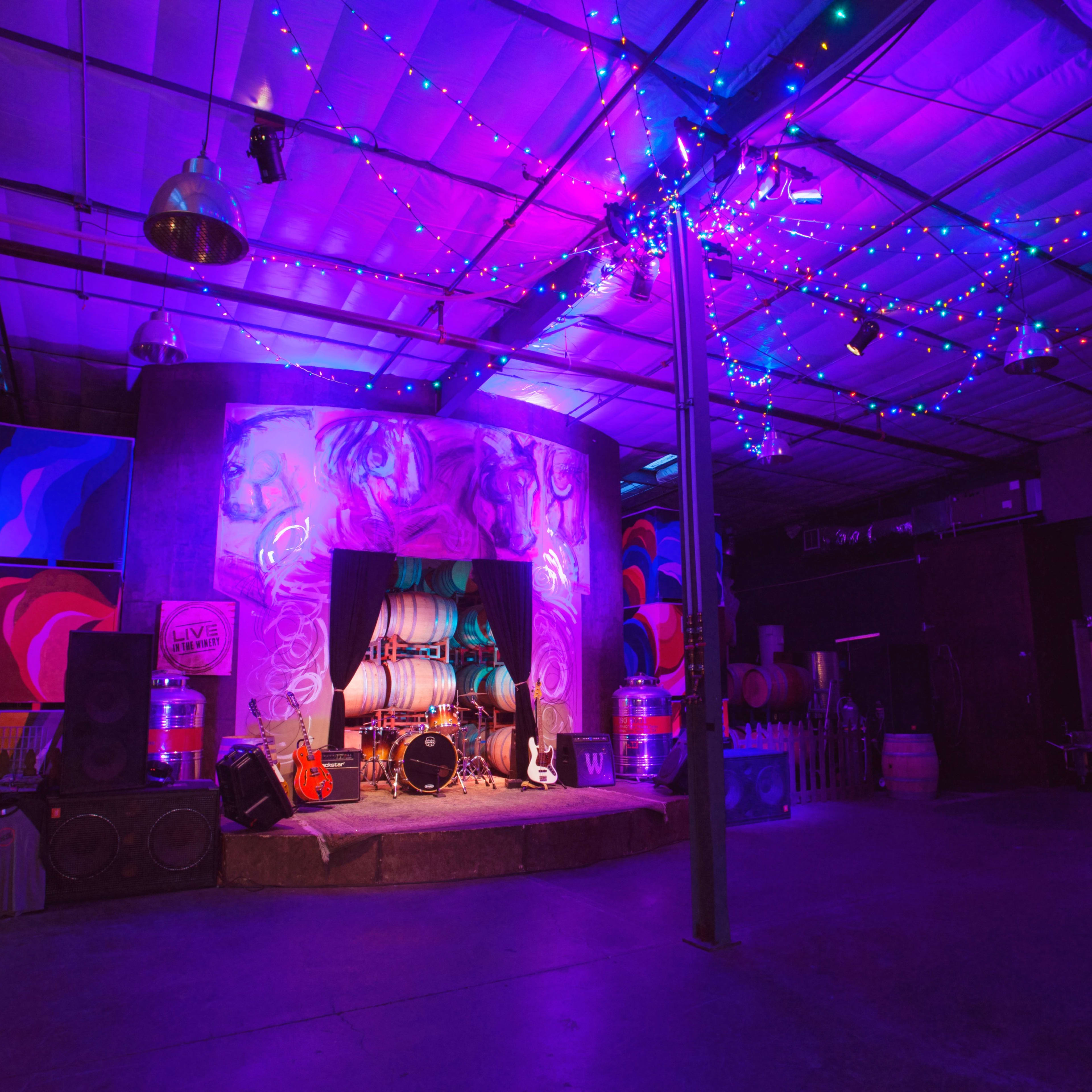 A dimly lit event space with colorful lights, featuring a decorated stage, musical instruments, and wine barrels in the background.