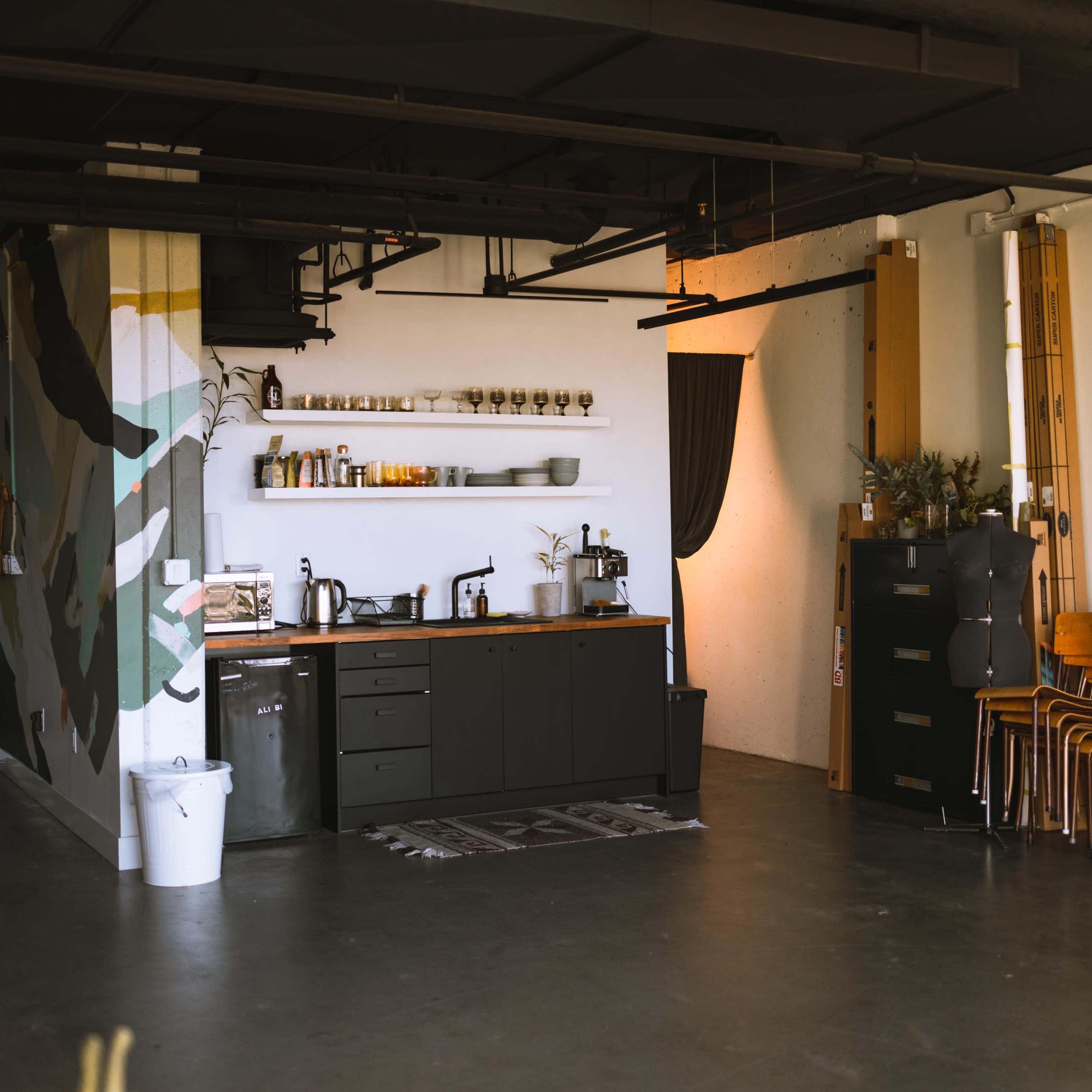 A modern kitchen area with a wooden counter, shelves of glassware, and a black cabinet against a wall featuring abstract art.