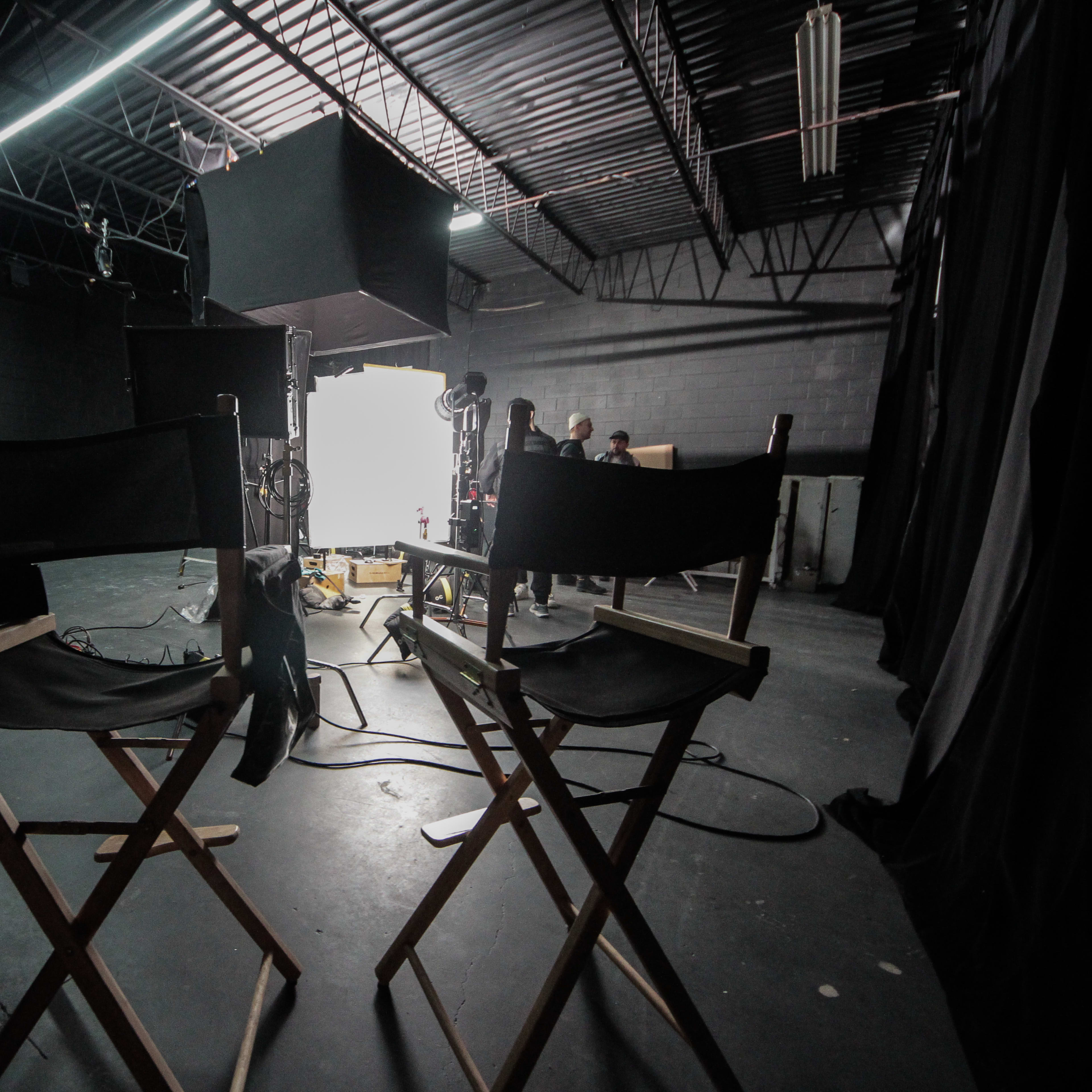Two director chairs face a bright light source in a dimly lit studio space, with equipment and crew visible in the background.