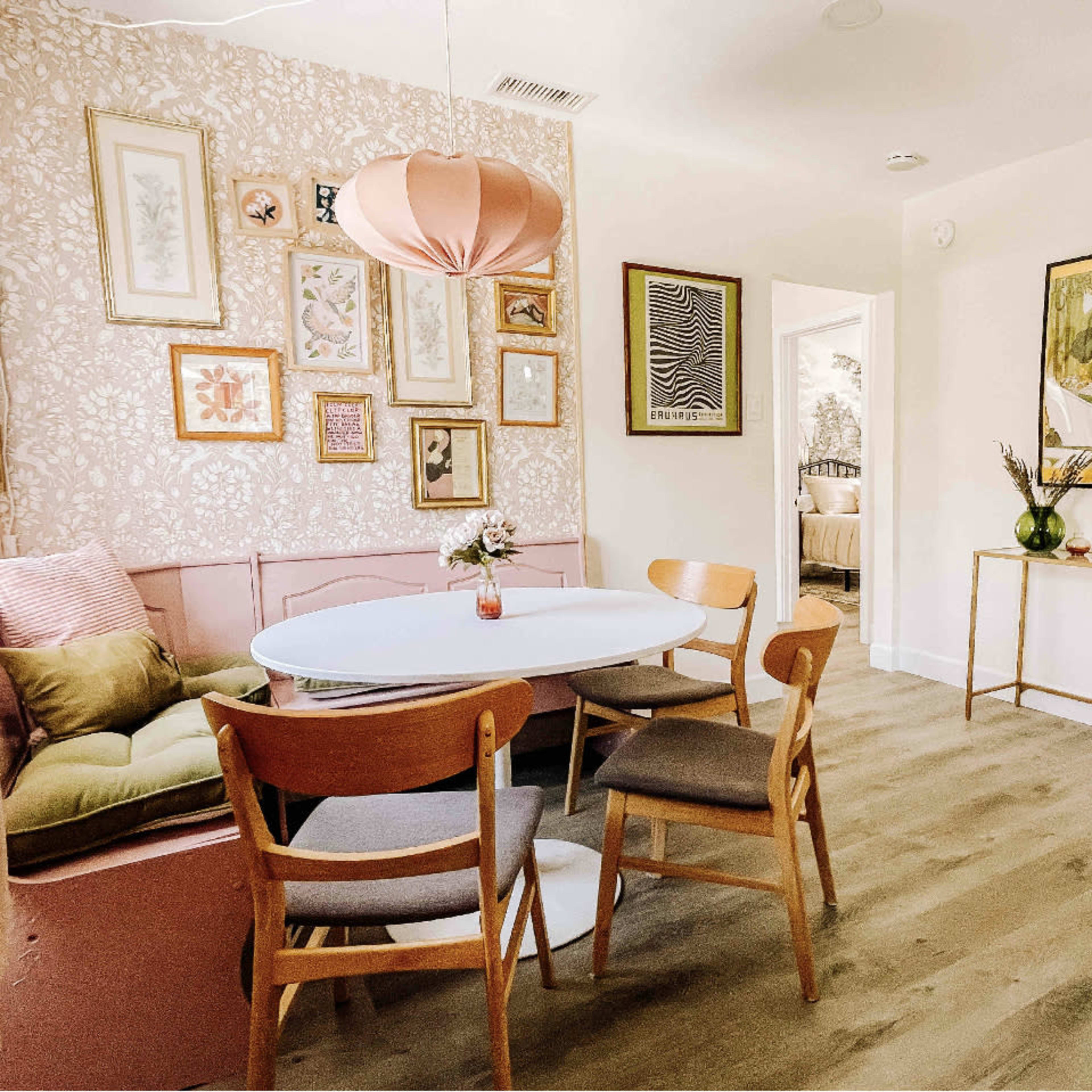 A dining area features a round table surrounded by wooden chairs, with a pink sofa against a floral wallpaper and framed artwork on the walls.