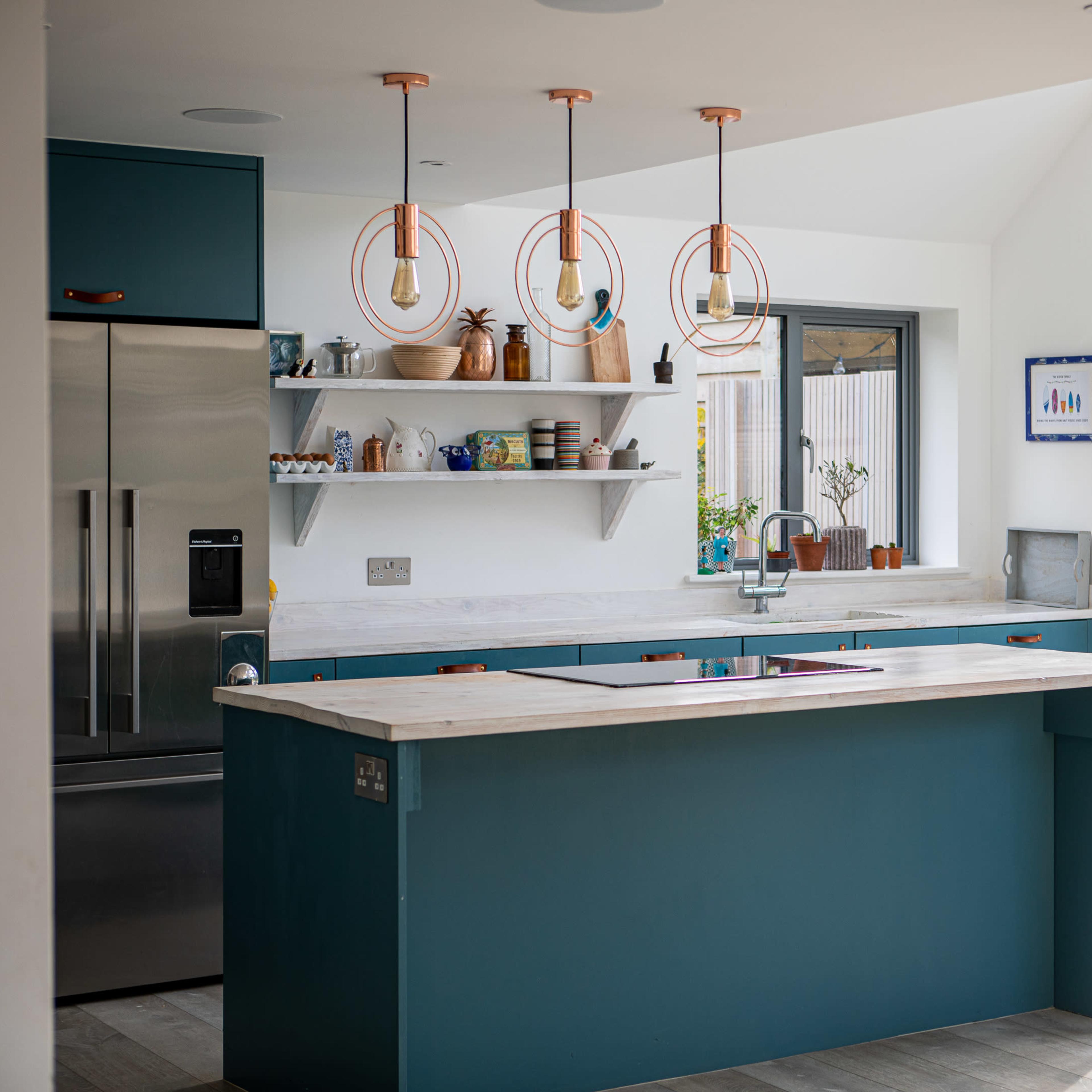 A modern kitchen with teal cabinetry, a central island, and pendant lights hanging above it, featuring a mixture of open shelves and stainless steel appliances.