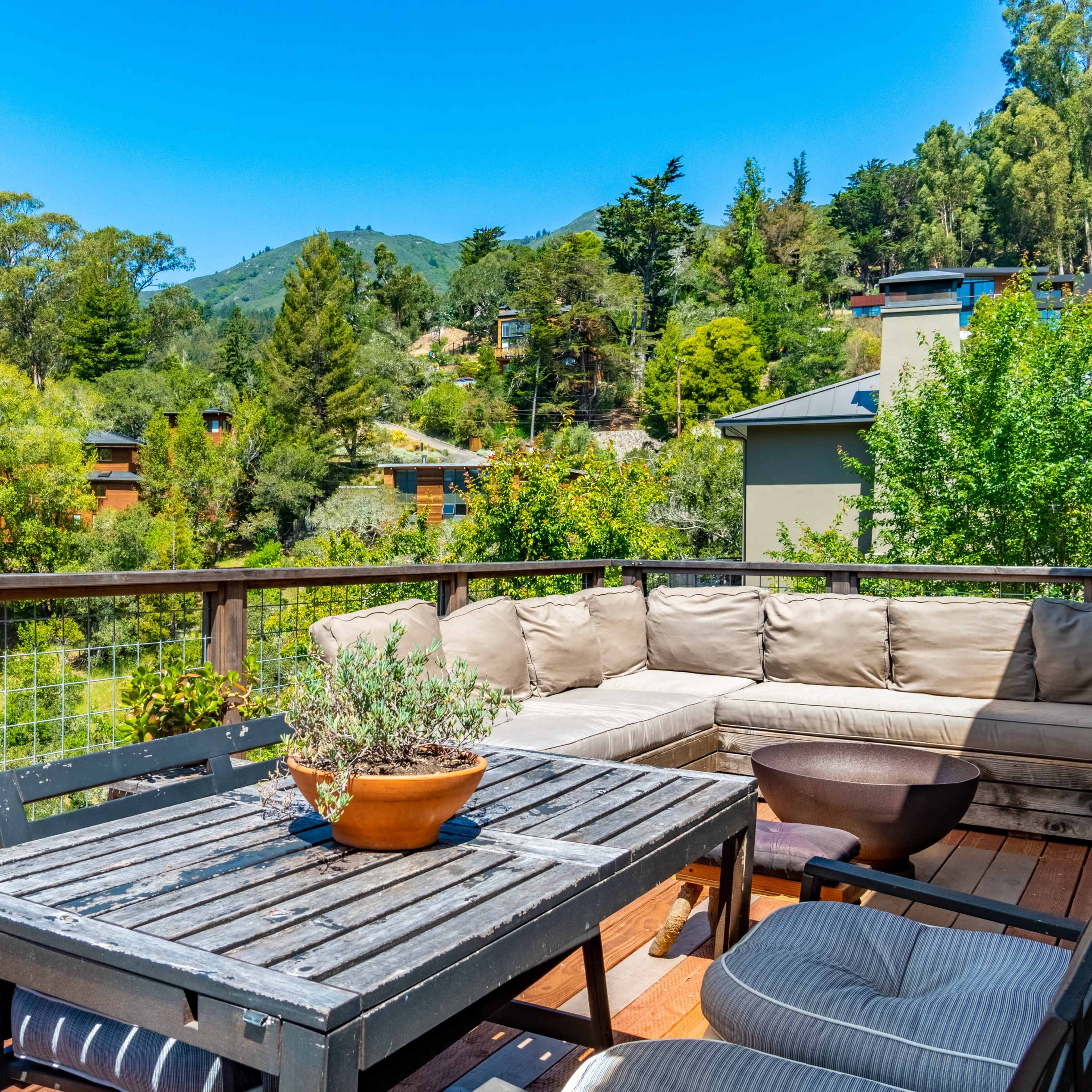 A wooden deck with a seating area and a table overlooks a green landscape with trees and houses.