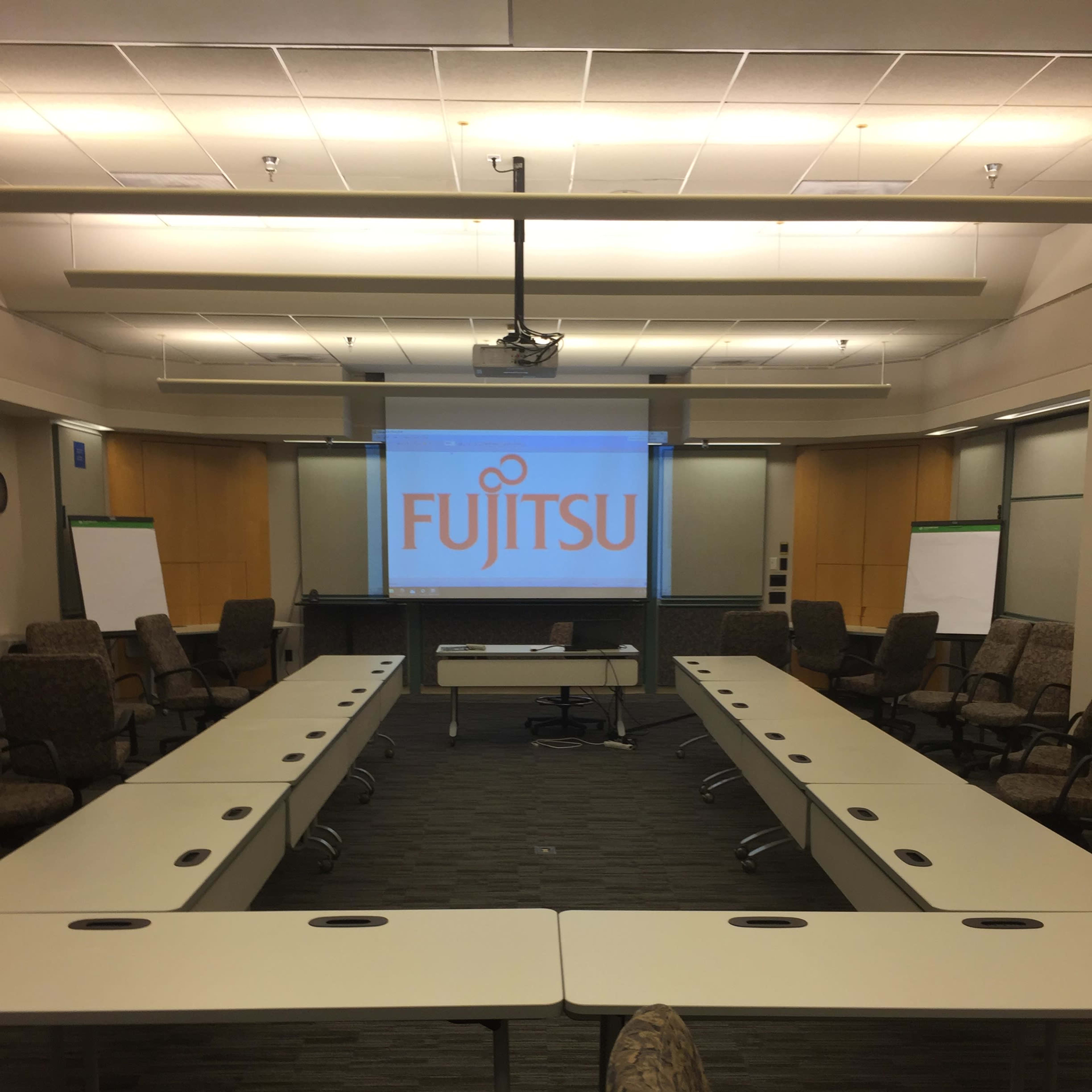A conference room is organized with tables arranged in a U-shape, featuring a projector screen displaying the Fujitsu logo at the front.