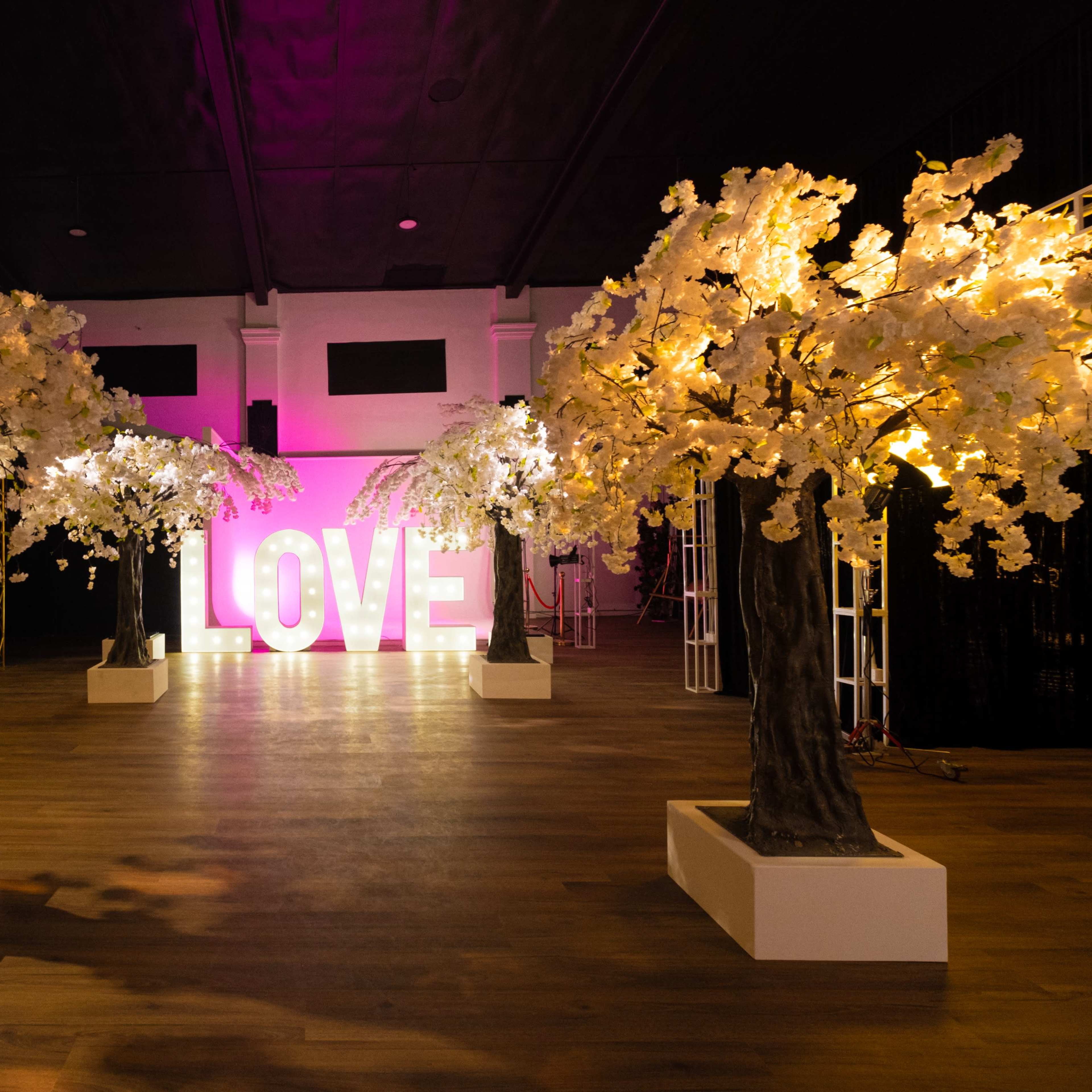 The scene features large illuminated letters spelling "LOVE" flanked by artificial cherry blossom trees in a dimly lit venue.