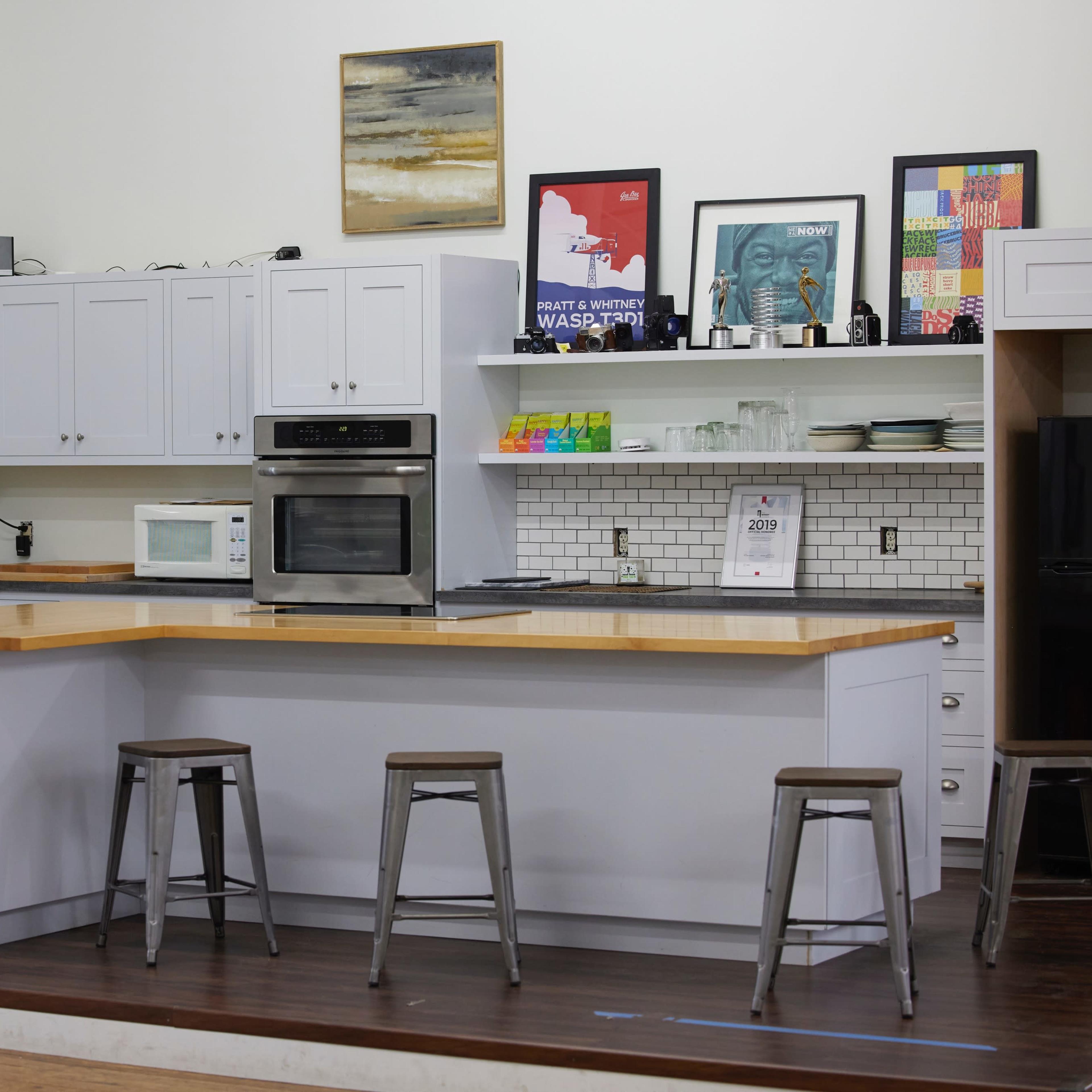 A modern kitchen features a central wooden island with four metal stools, white cabinets, and various appliances including a microwave and oven.