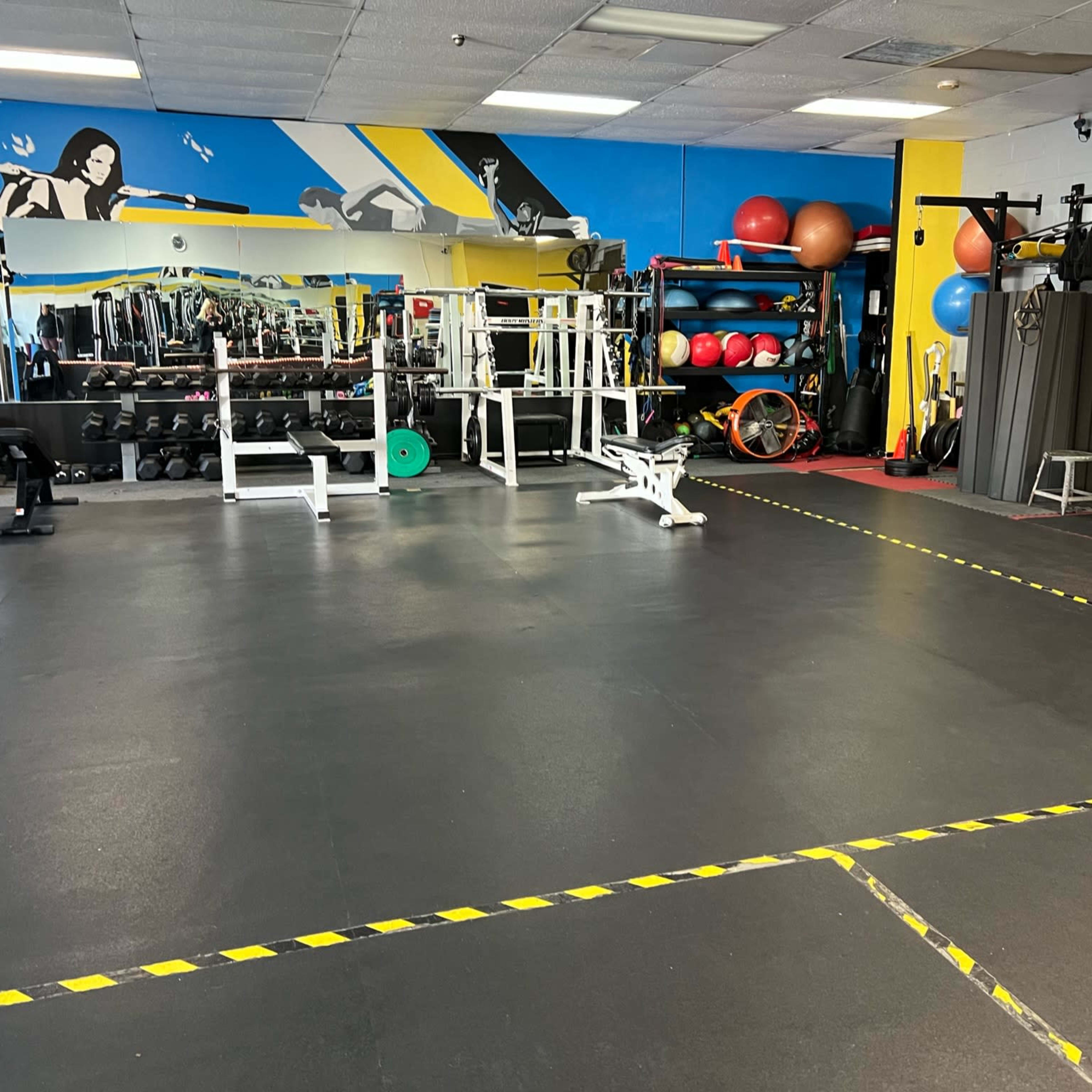 A gym interior with various exercise equipment, including weight benches, dumbbells, and fitness balls, against a colorful mural.