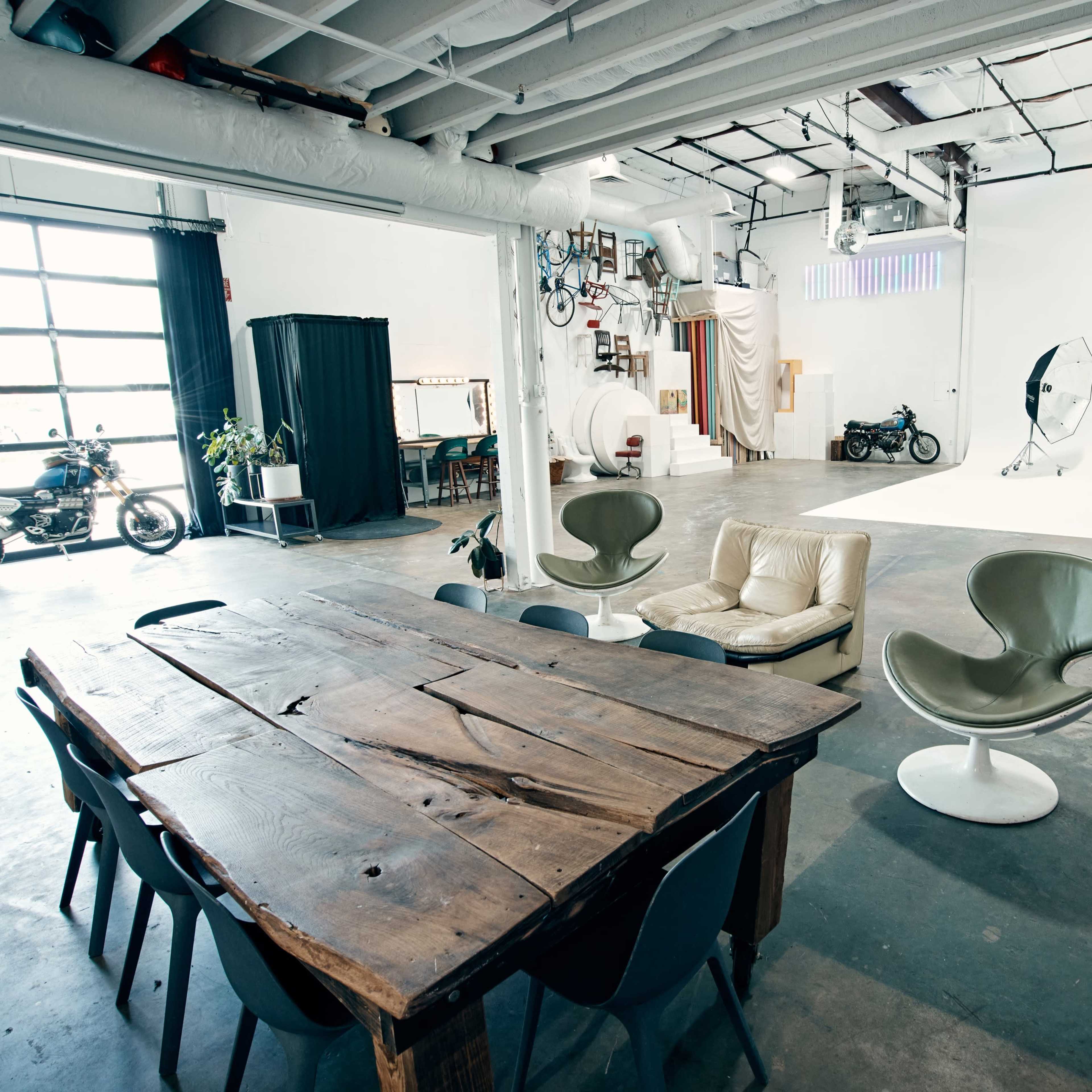A spacious studio with a wooden dining table, multiple seating areas, and a large white backdrop for photography, alongside a motorcycle and decor on the walls.