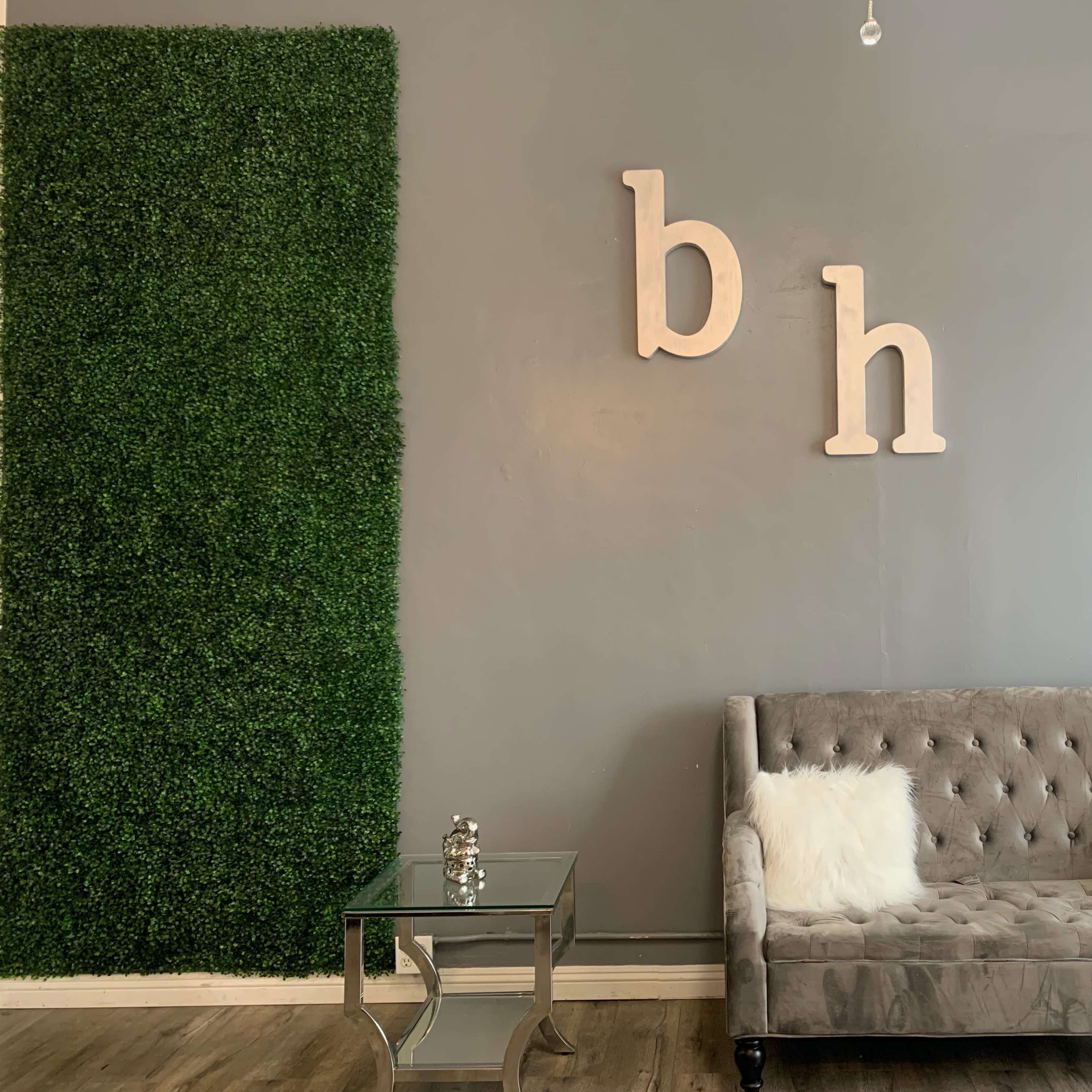 A gray wall with decorative greenery on one side, a tufted gray sofa, and a glass side table with a decorative item on it.