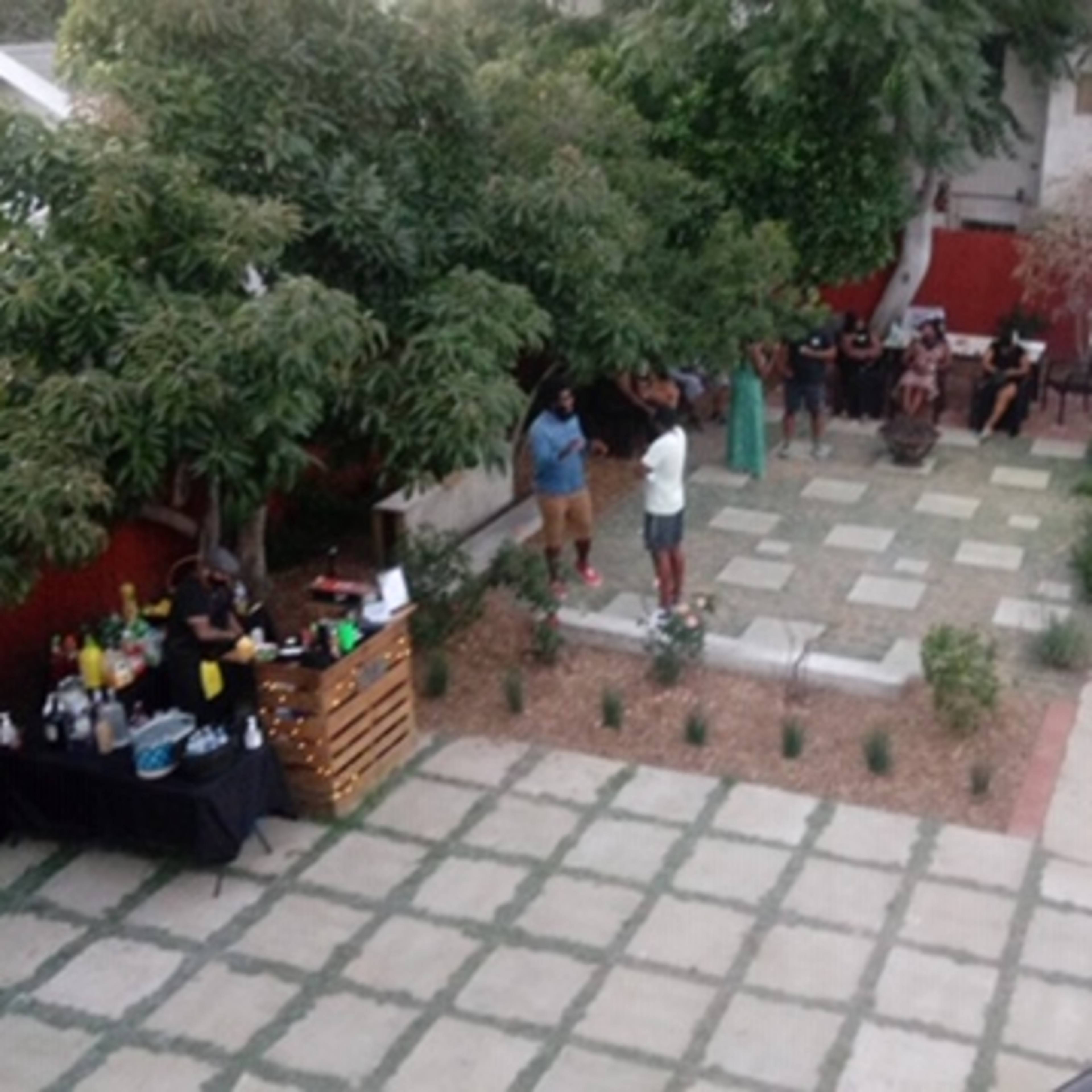 A gathering is taking place in a backyard with people socializing around a stone-paved area, while food and drinks are set up on tables nearby.