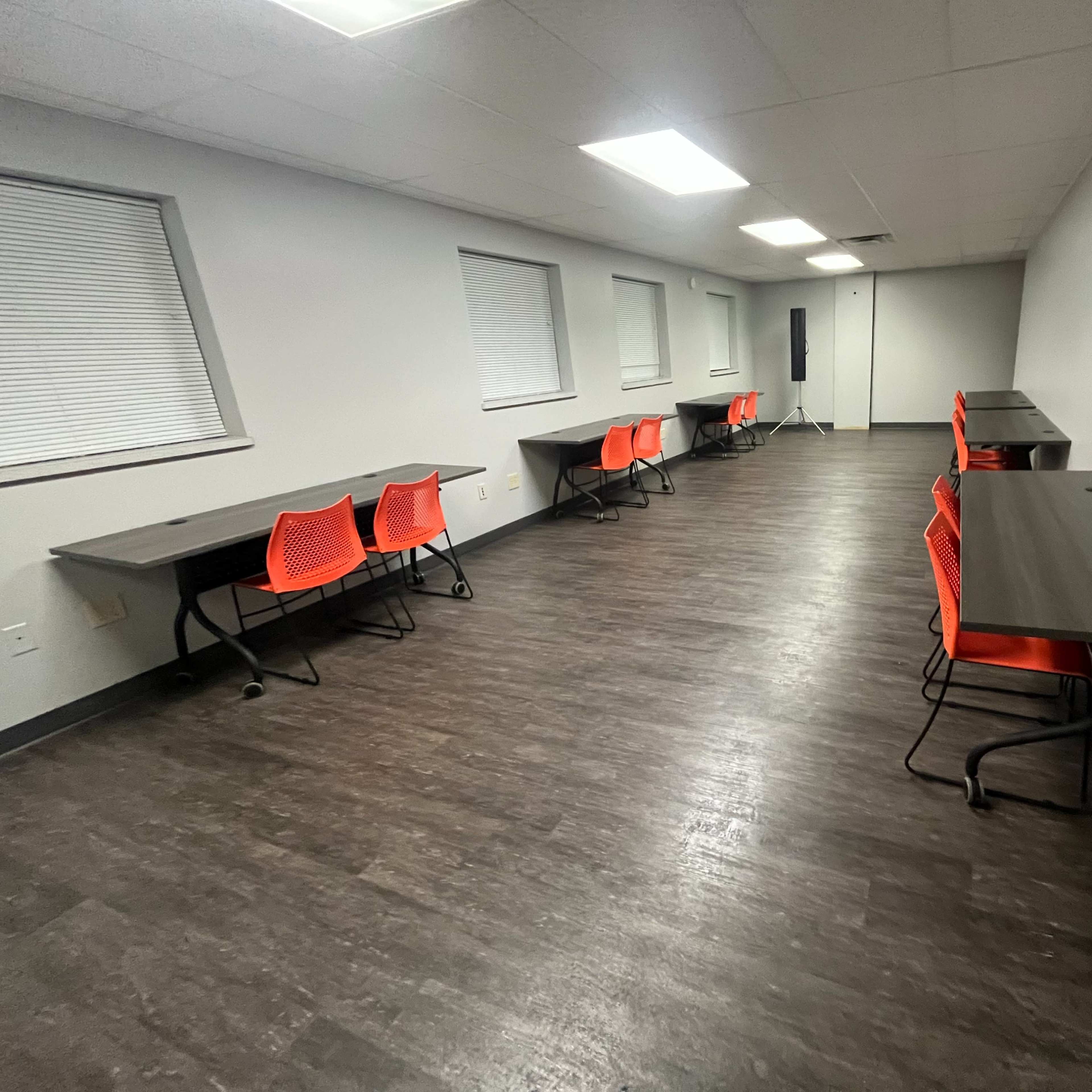 A room with five tables and orange chairs arranged in two parallel rows along the walls, with windows on the left side and gray flooring.