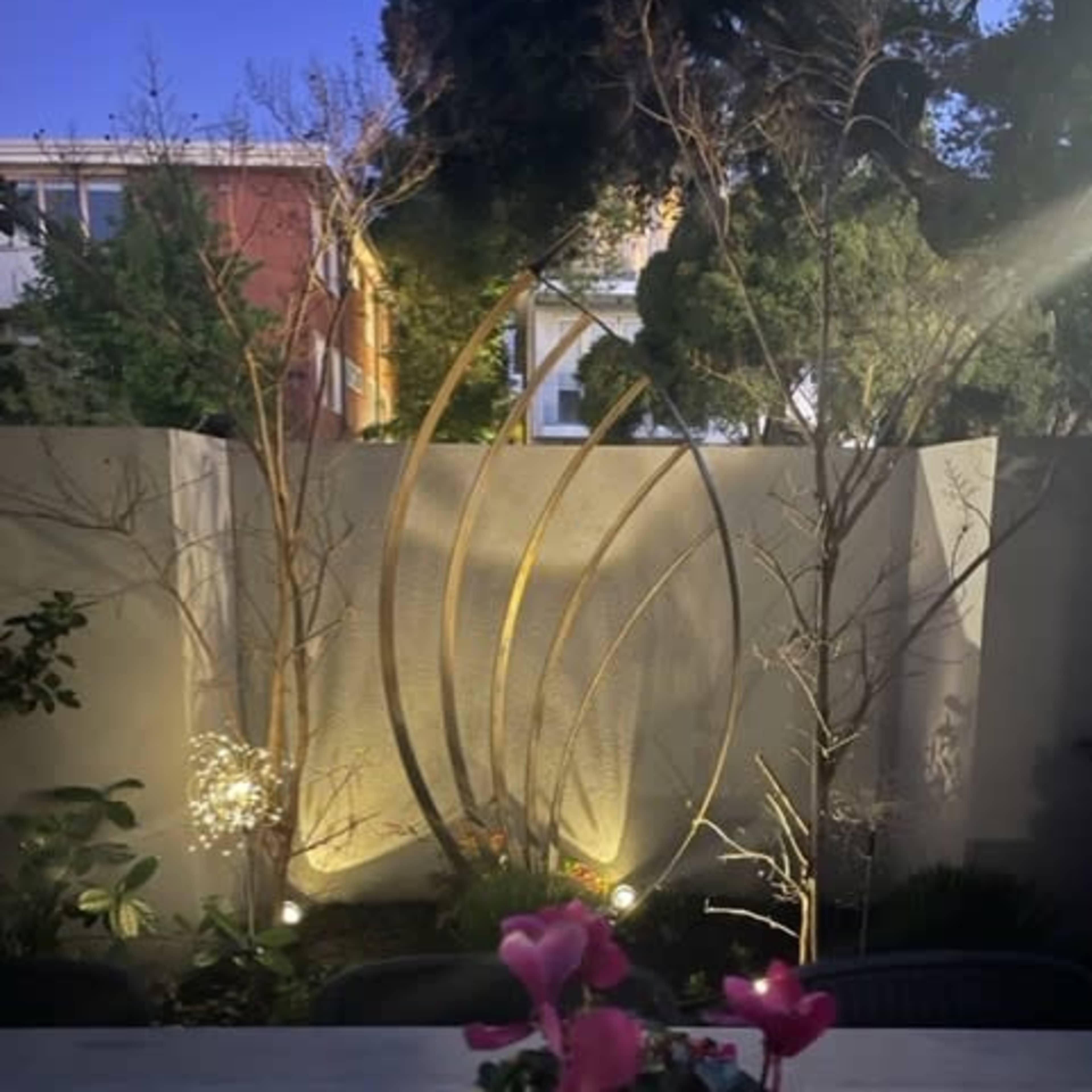 A potted plant with pink flowers sits on a wooden table in the foreground, while a softly lit garden scene with decorative metal sculptures and trees is visible in the background.