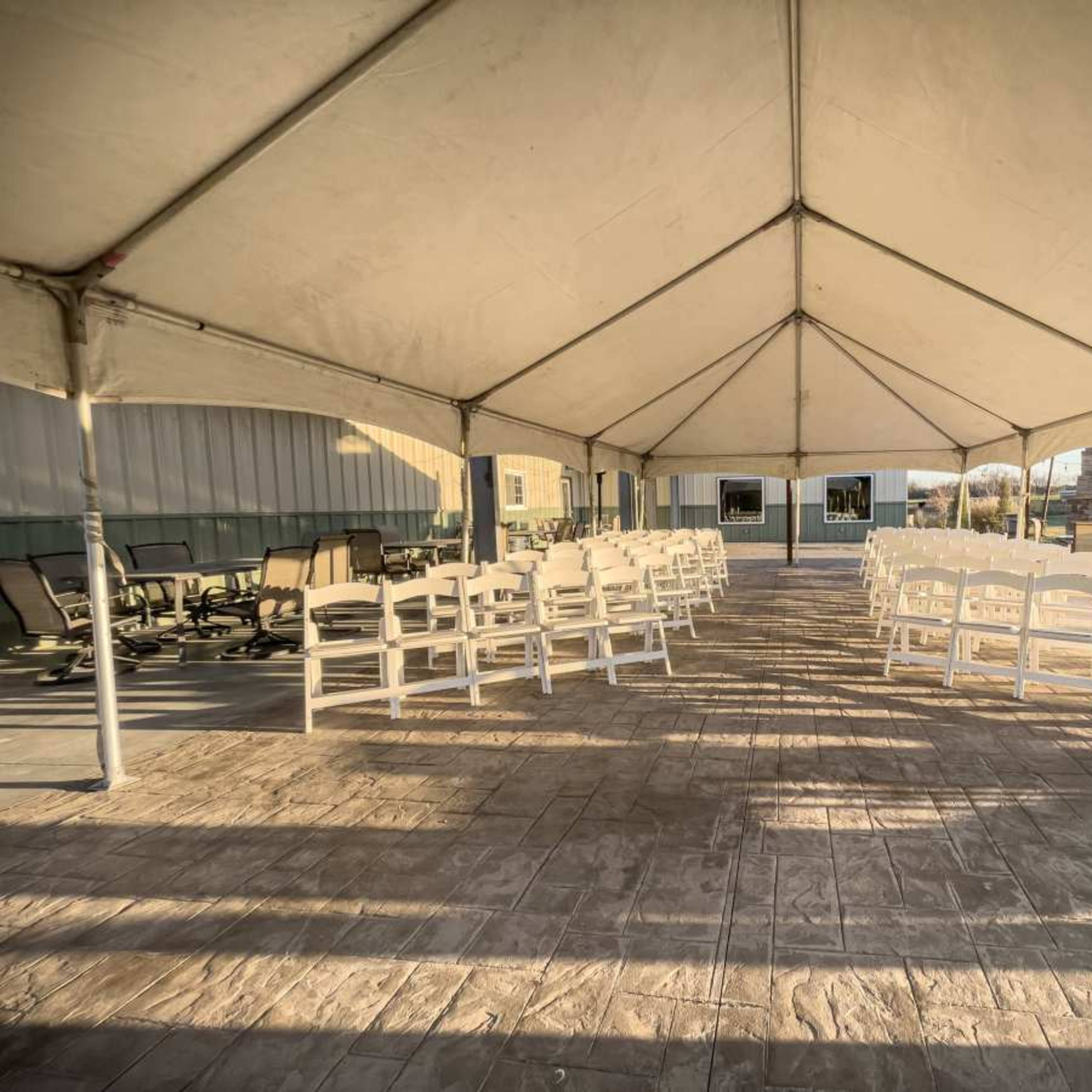 A large tent is set up with rows of white chairs arranged on a concrete surface, leading towards a building in the background.