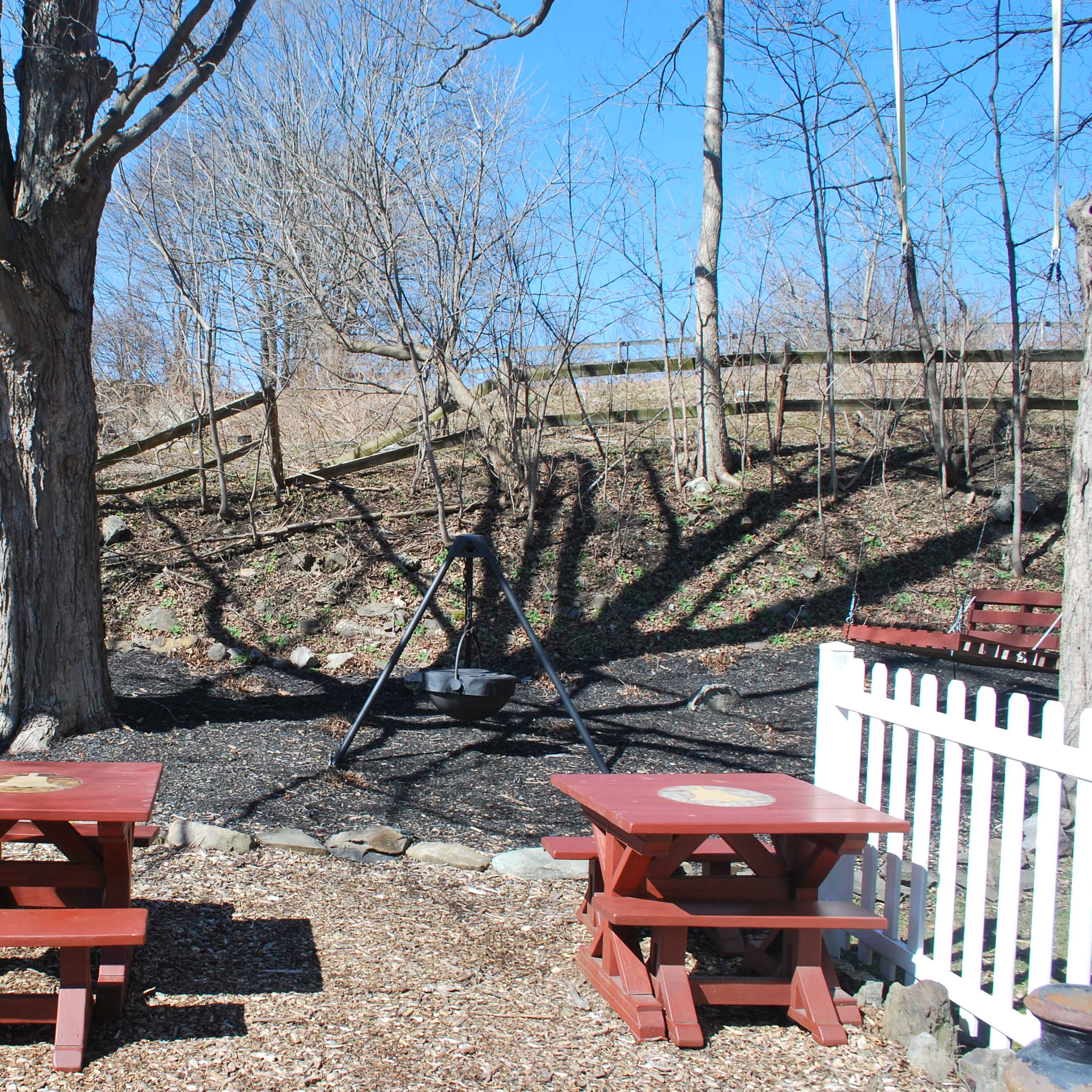A playground area with two red picnic tables and a swing set in a yard featuring bare trees and a sloped, mulched terrain.