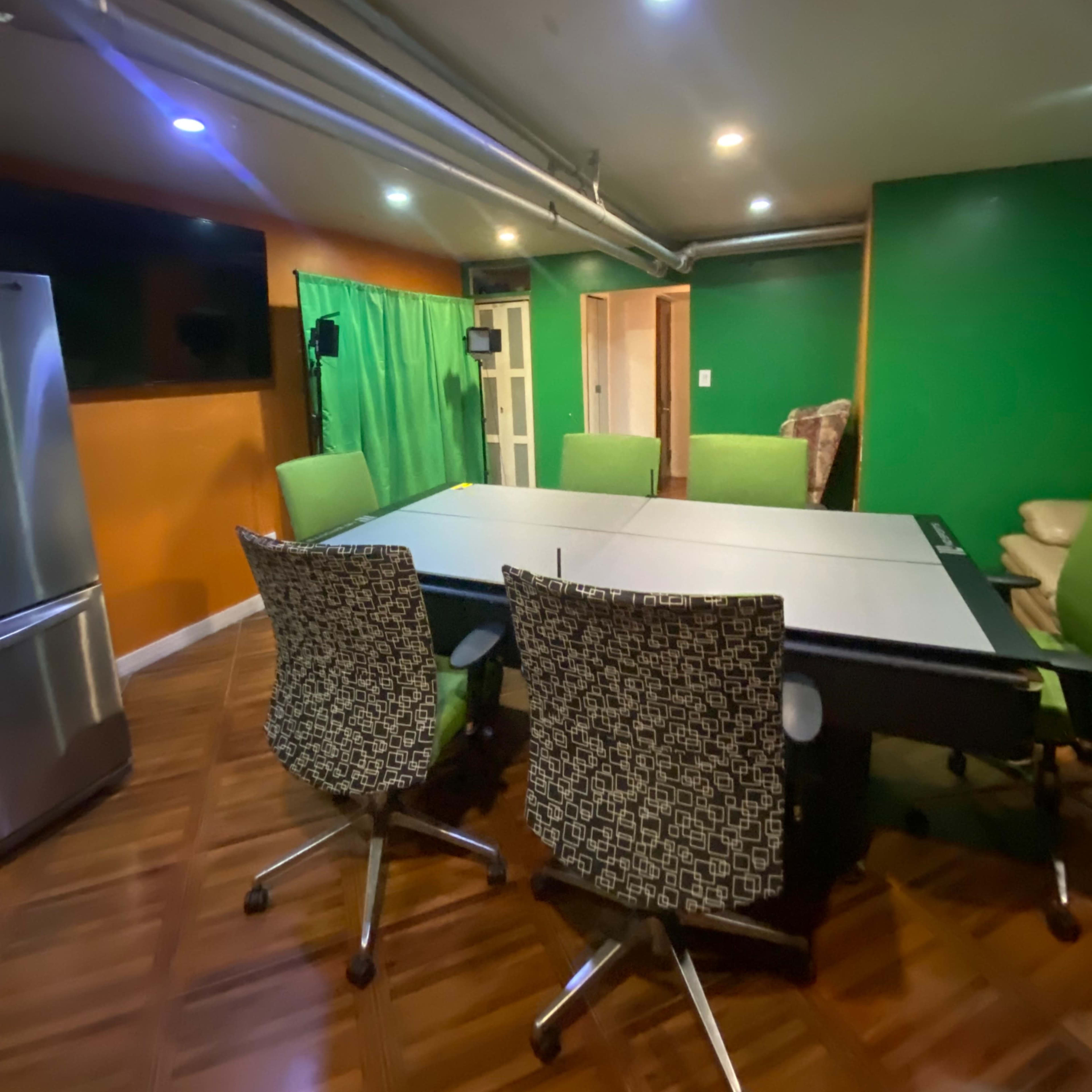 A modern conference room with a large table surrounded by ergonomic chairs, green walls, and a fridge in the corner.