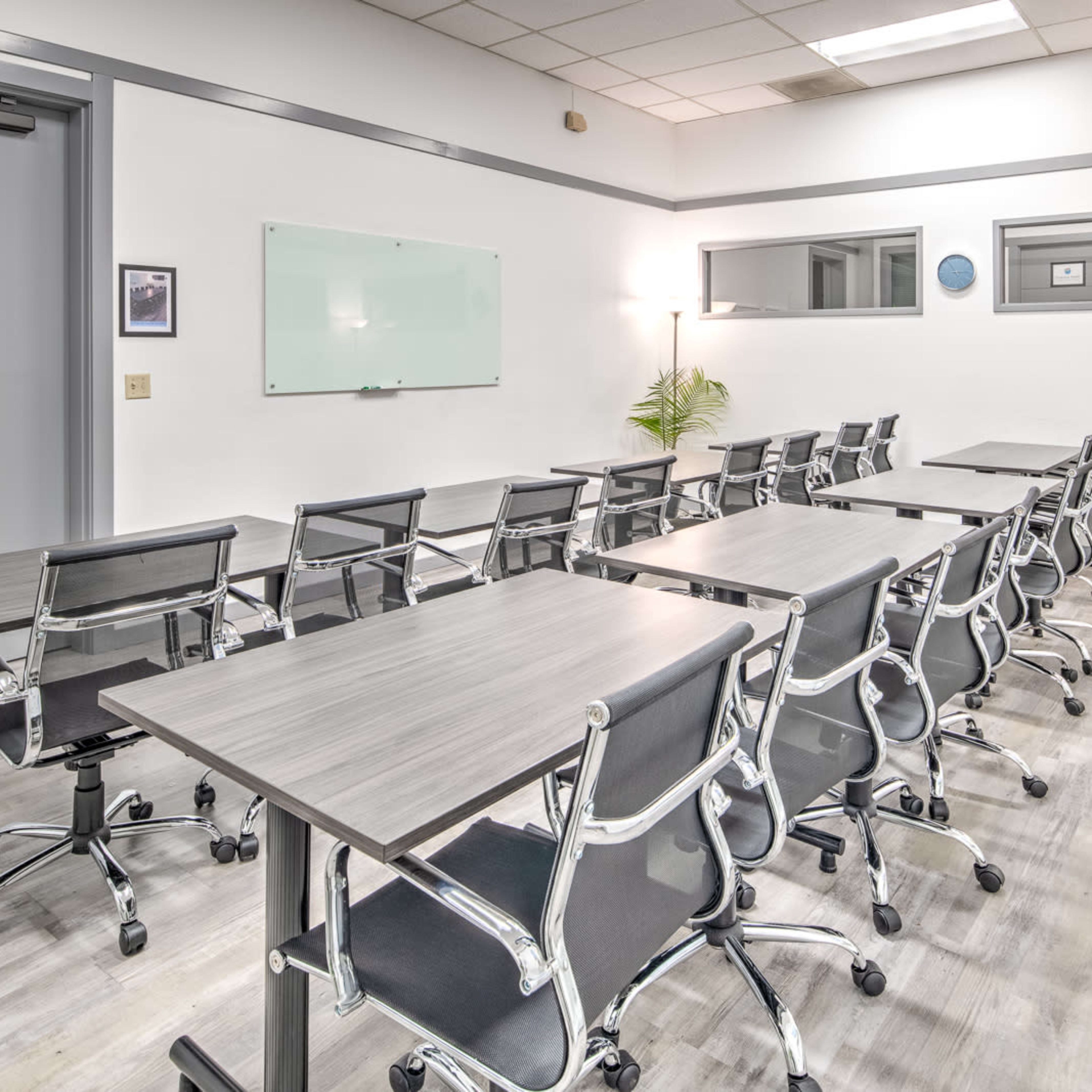 A modern conference room with several rows of tables and chairs, equipped with a whiteboard and plants.