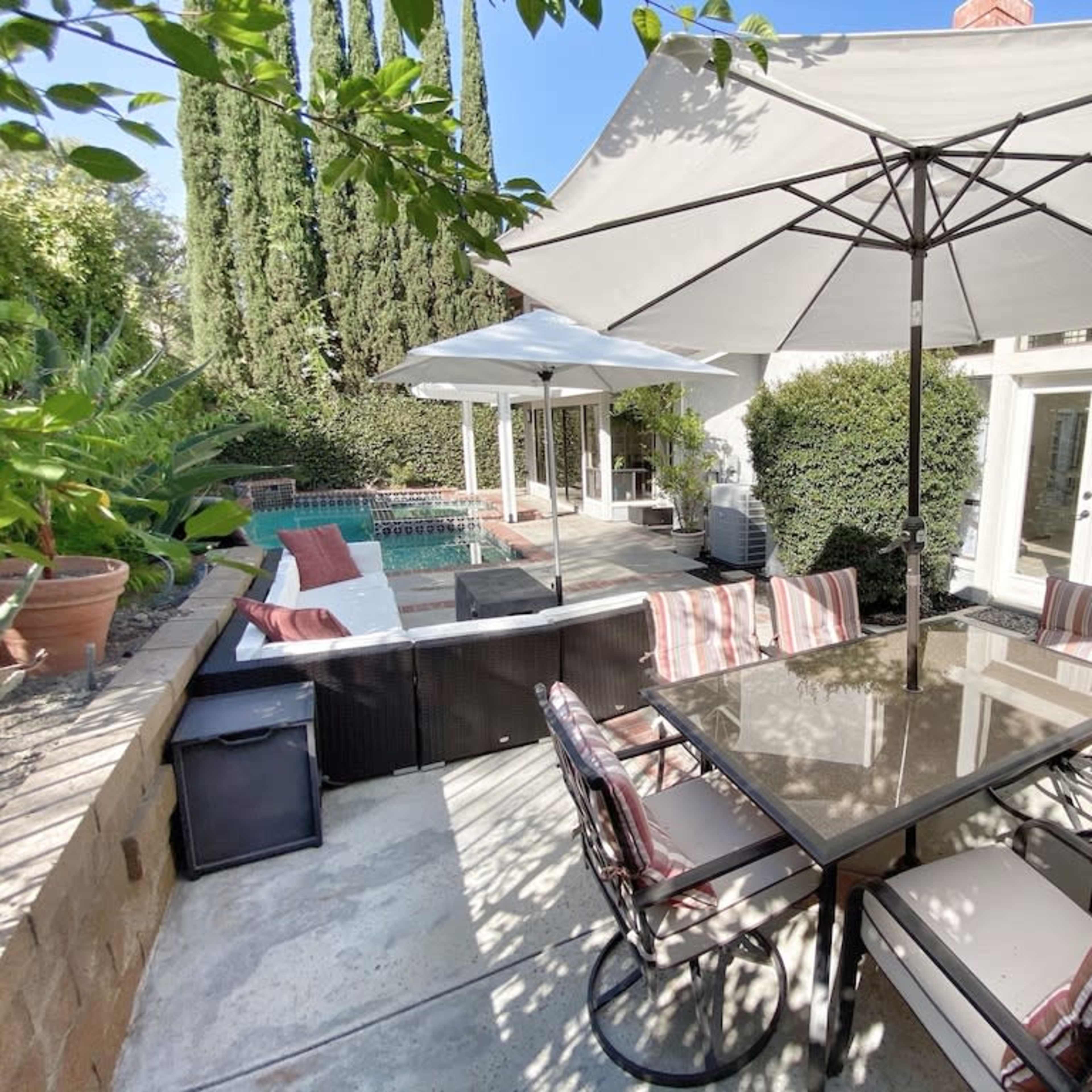 A patio area features a table with several chairs, a swimming pool, and umbrellas providing shade in a landscaped backyard.