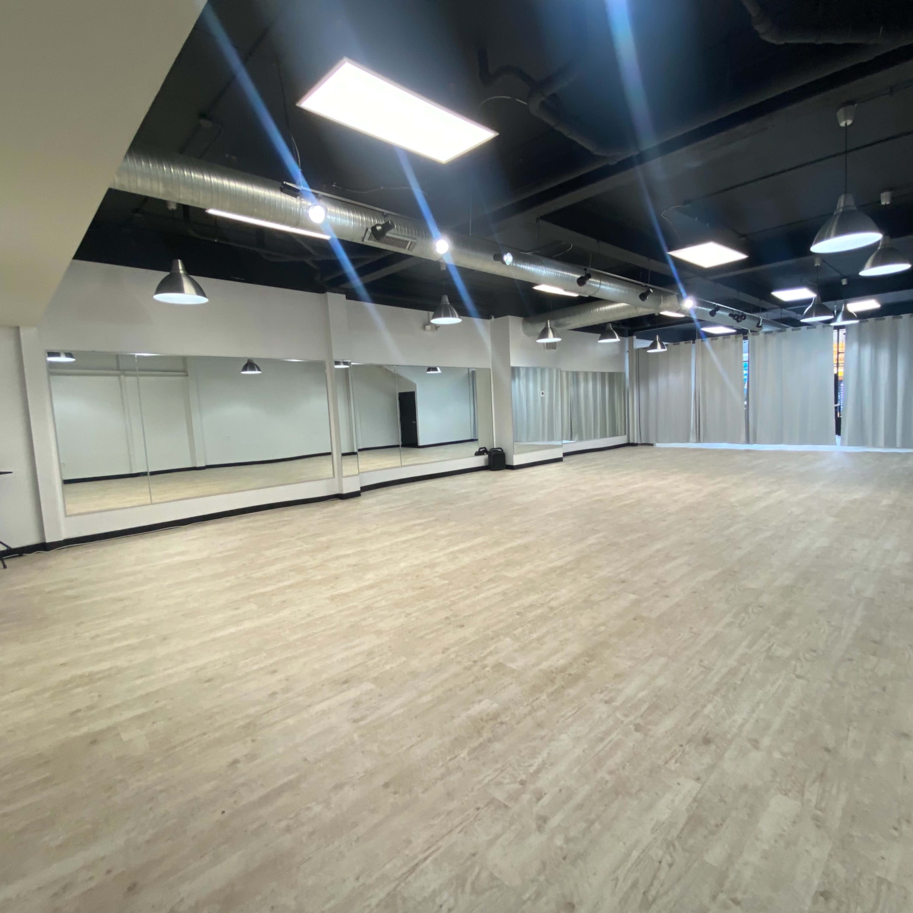 A spacious studio with a light wooden floor, mirrored walls, and overhead lights.