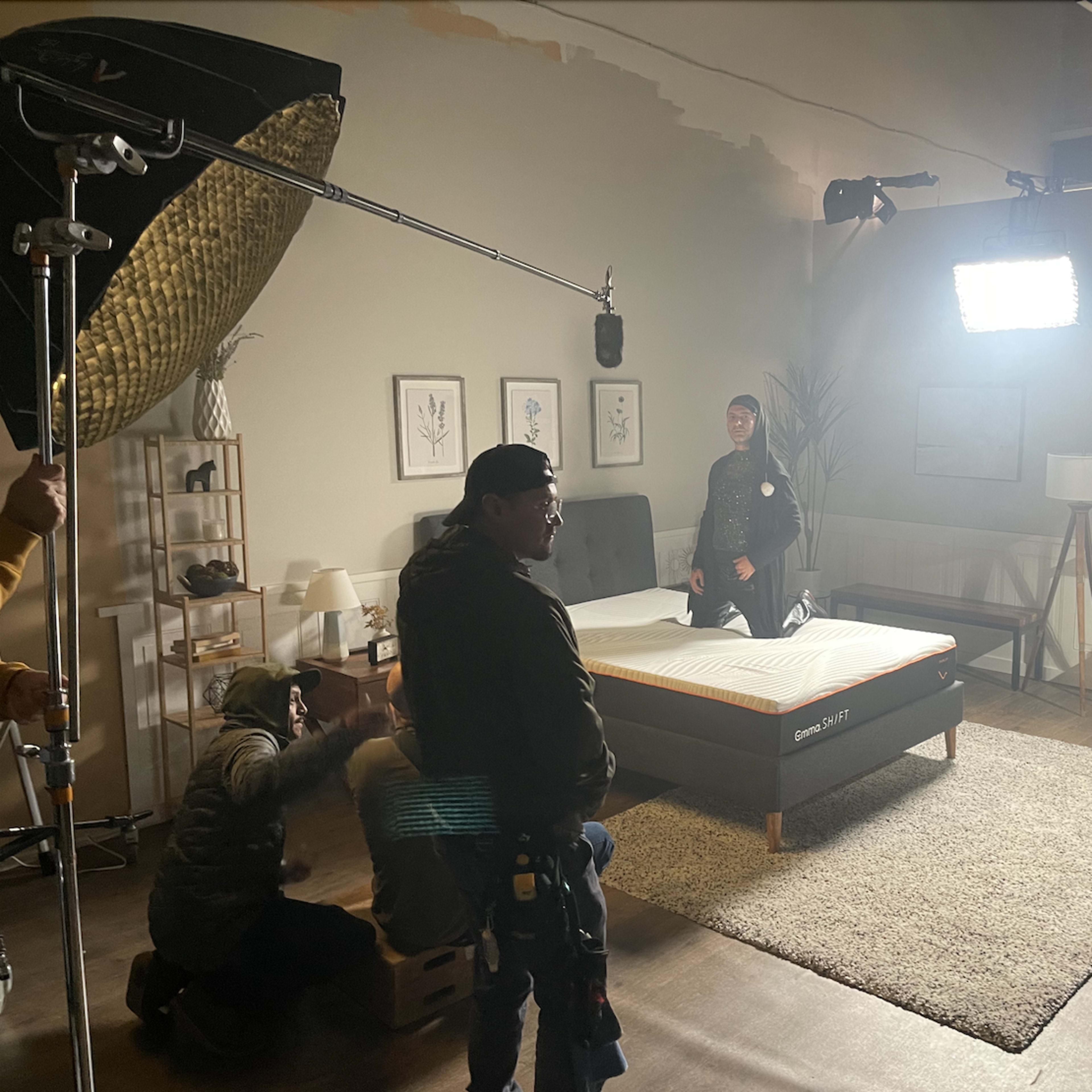 A film crew is setting up shots in a room featuring a bed, with lights and microphones arranged around the scene.