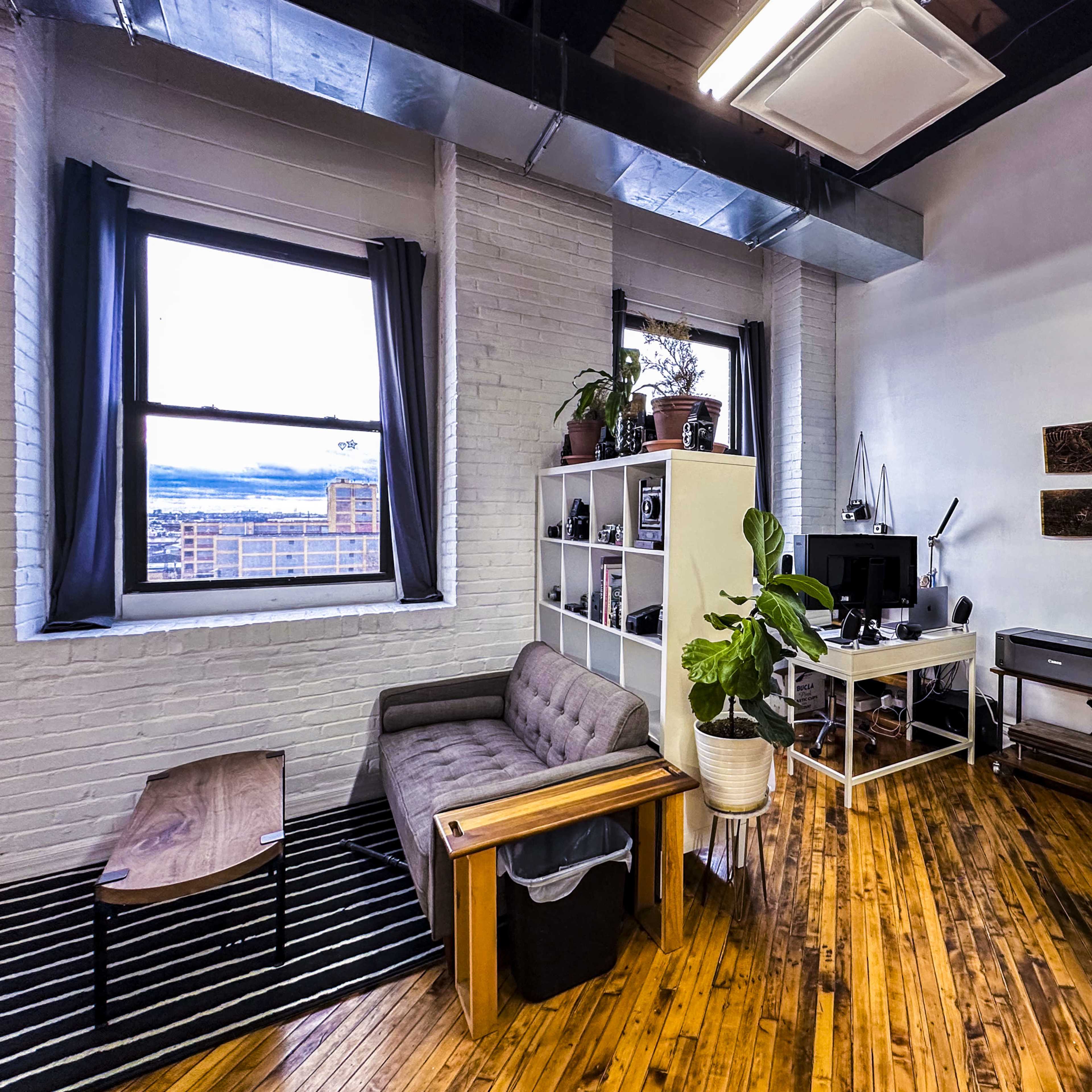 A cozy urban living space with a brown couch, a coffee table, a bookshelf, and a desk with a computer, all set against a backdrop of exposed brick walls and large windows.